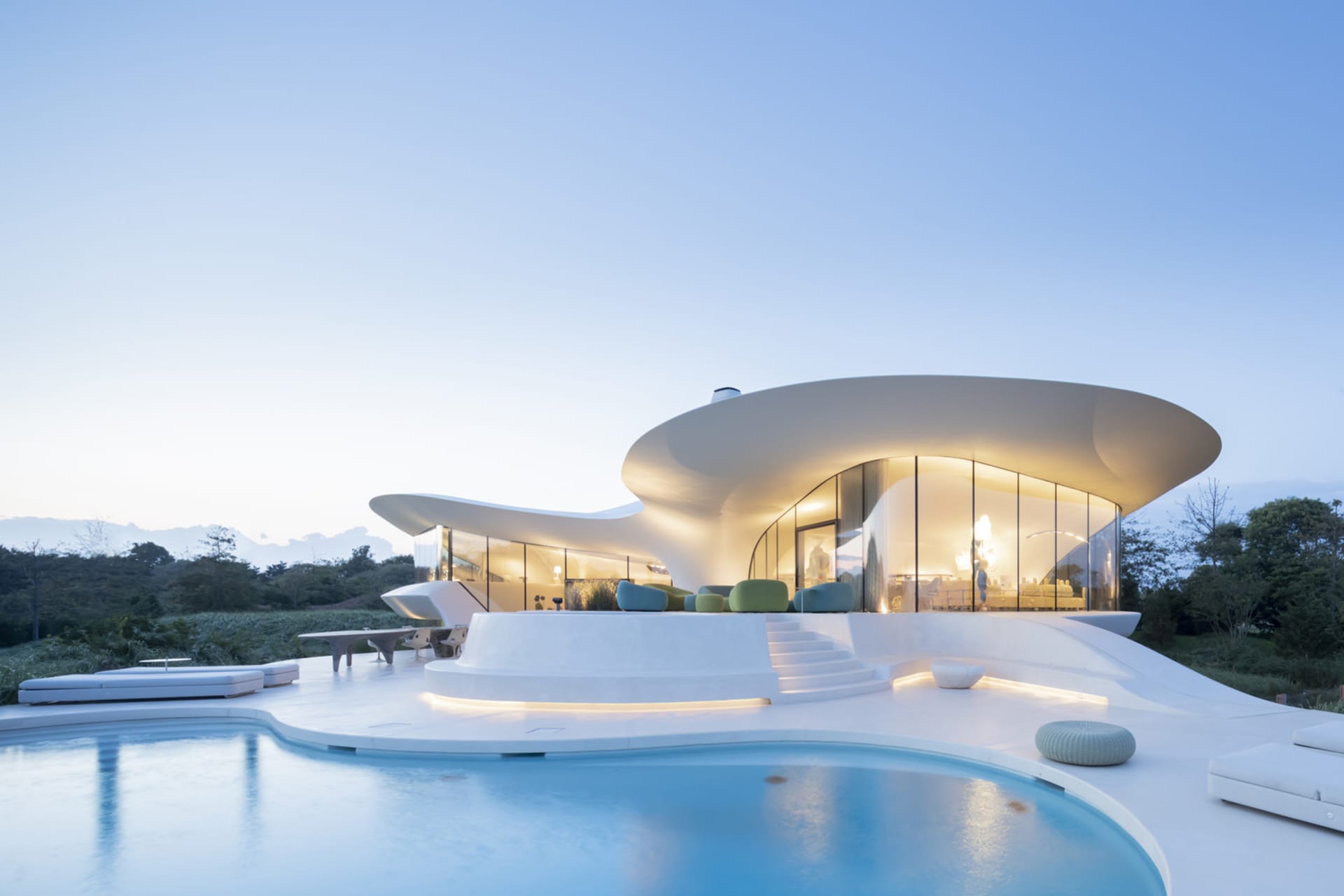 A modern house featuring large glass walls and a concrete structure, with a pool and outdoor seating area.