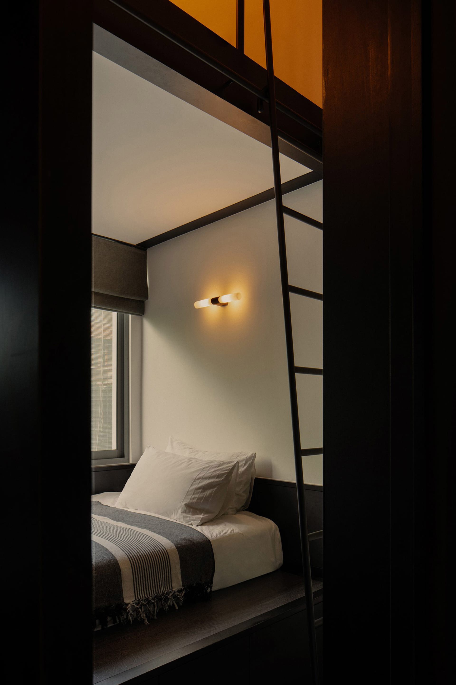 A minimalist bedroom with a single bed, pillows, and a blanket, featuring a wooden beam ceiling and a ladder.