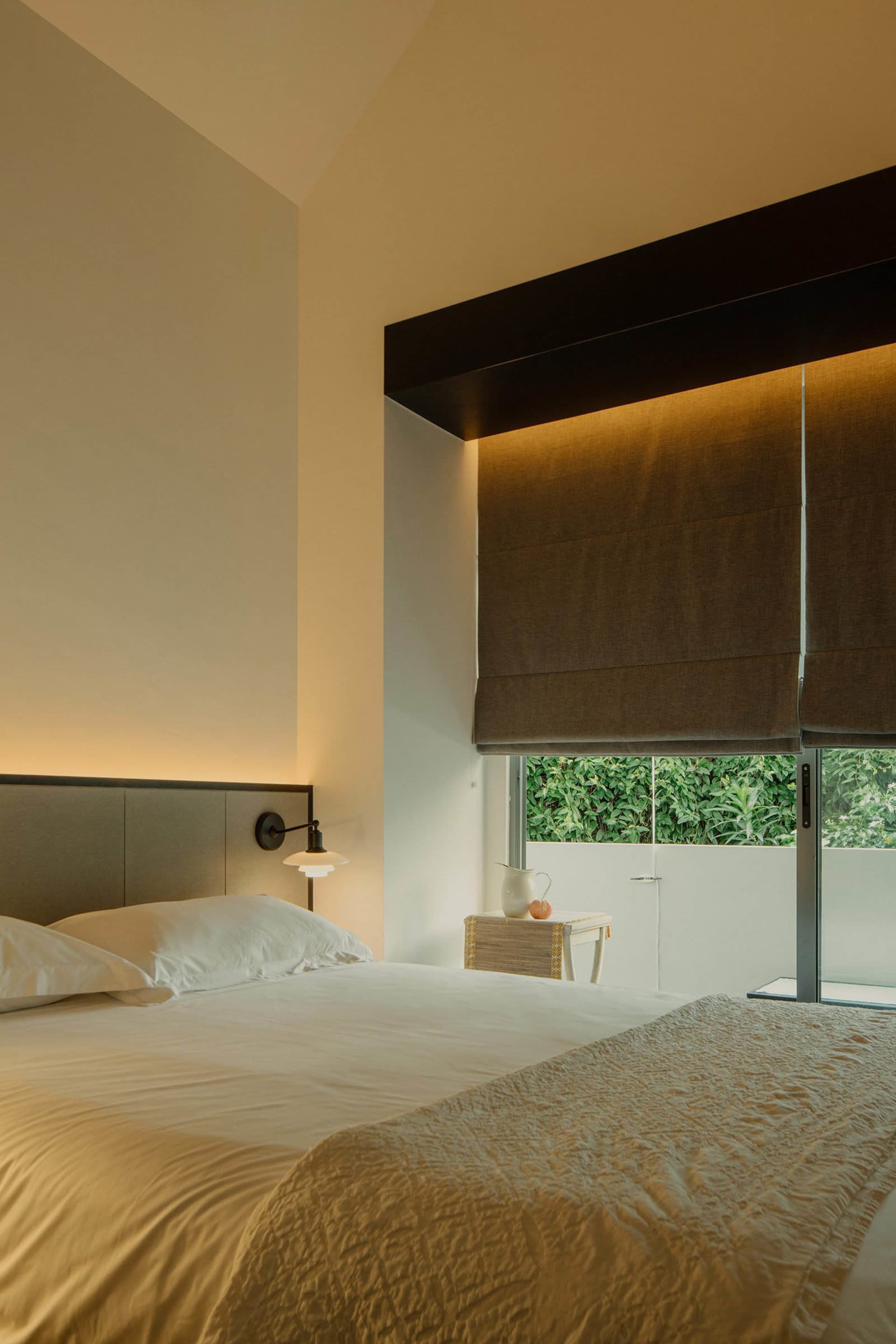 A minimalist bedroom with white walls, a textured fabric bedspread, and a window with dark blinds.