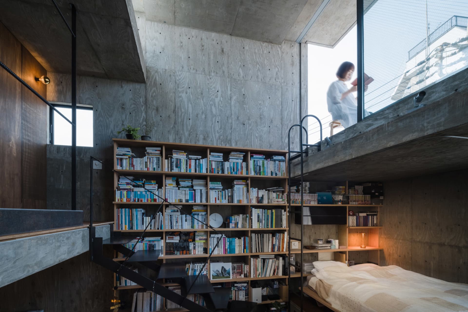A modern industrial bedroom featuring concrete walls, a wooden bookshelf filled with books, and a black metal staircase.