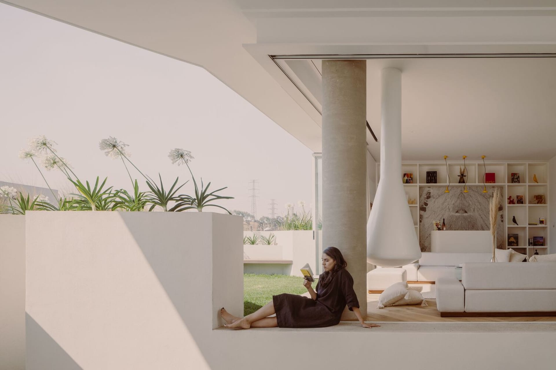 A minimalist living space with concrete walls and large windows, featuring a woman reading a book in a corner.