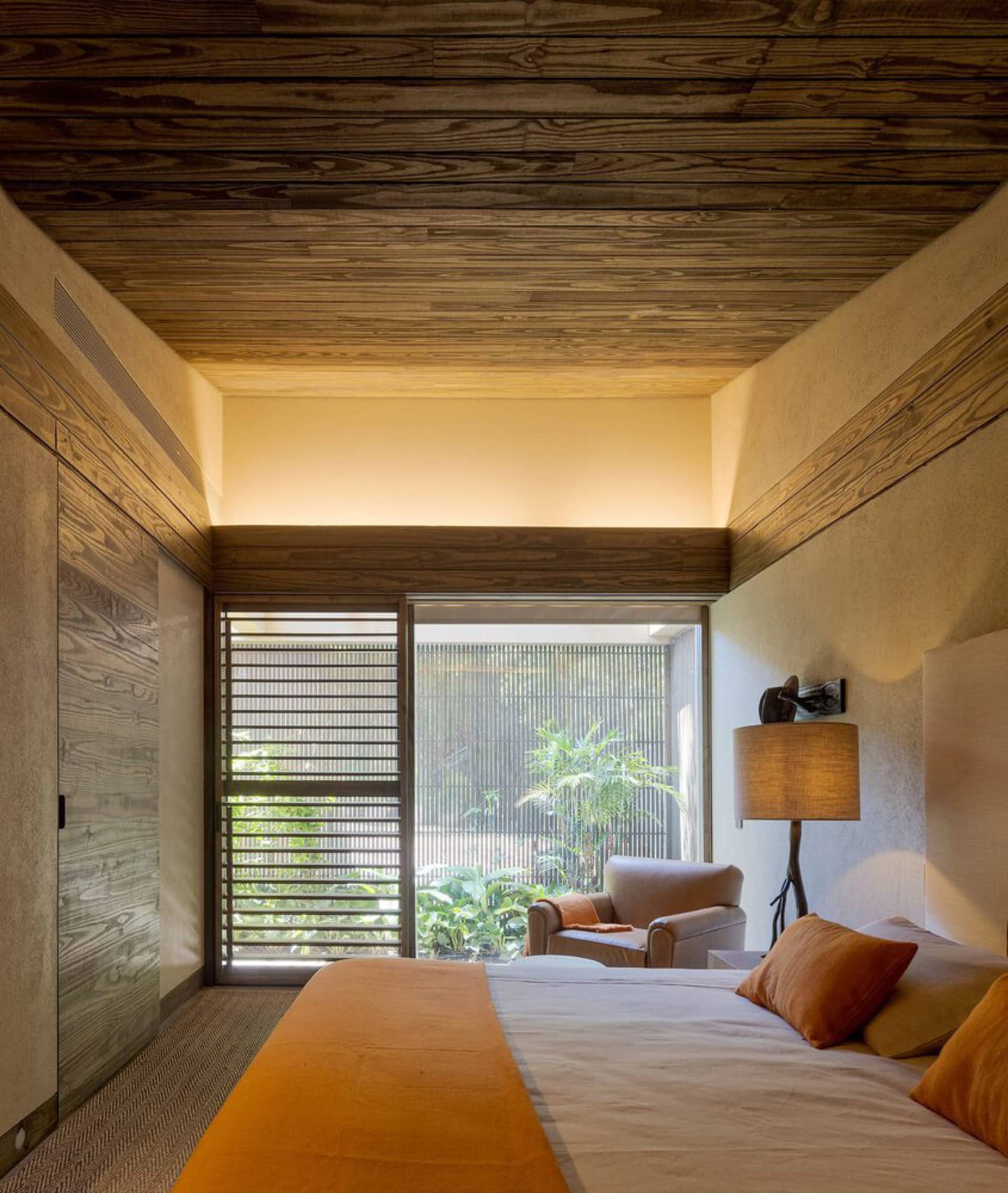 A modern bedroom with a wooden ceiling, orange bedding, and a view of a tropical garden.