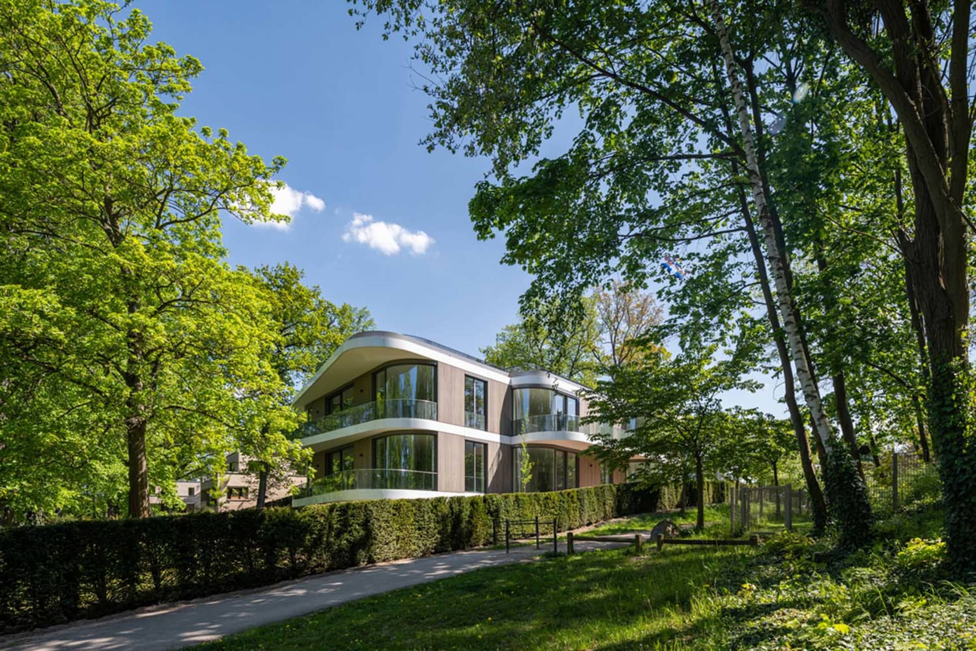 Modern residential house with wooden exterior and glass windows surrounded by greenery on a sunny day.