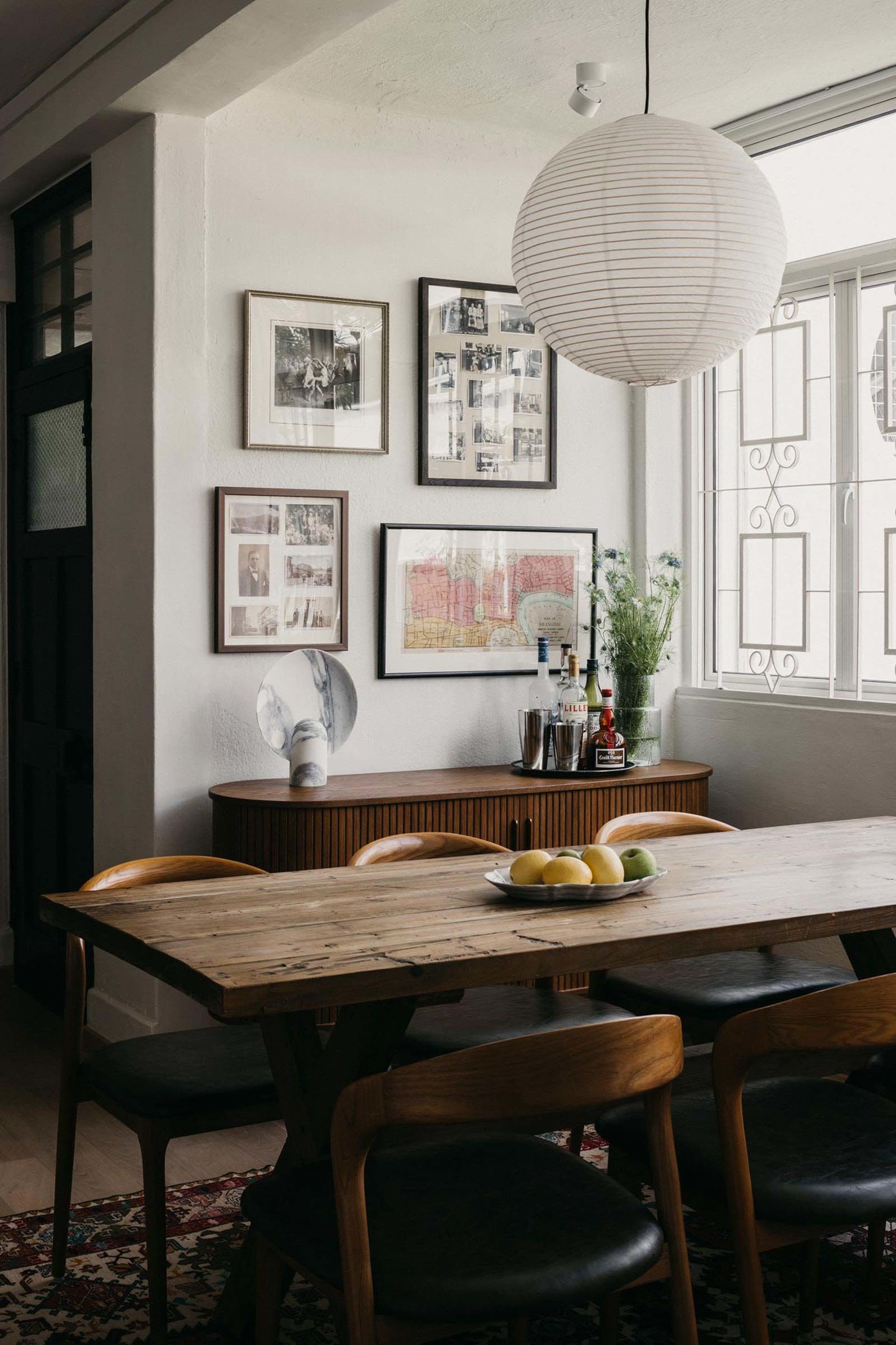 A modern dining room featuring a wooden table adorned with a bowl of fruit, a white pendant lamp, and framed photographs on t