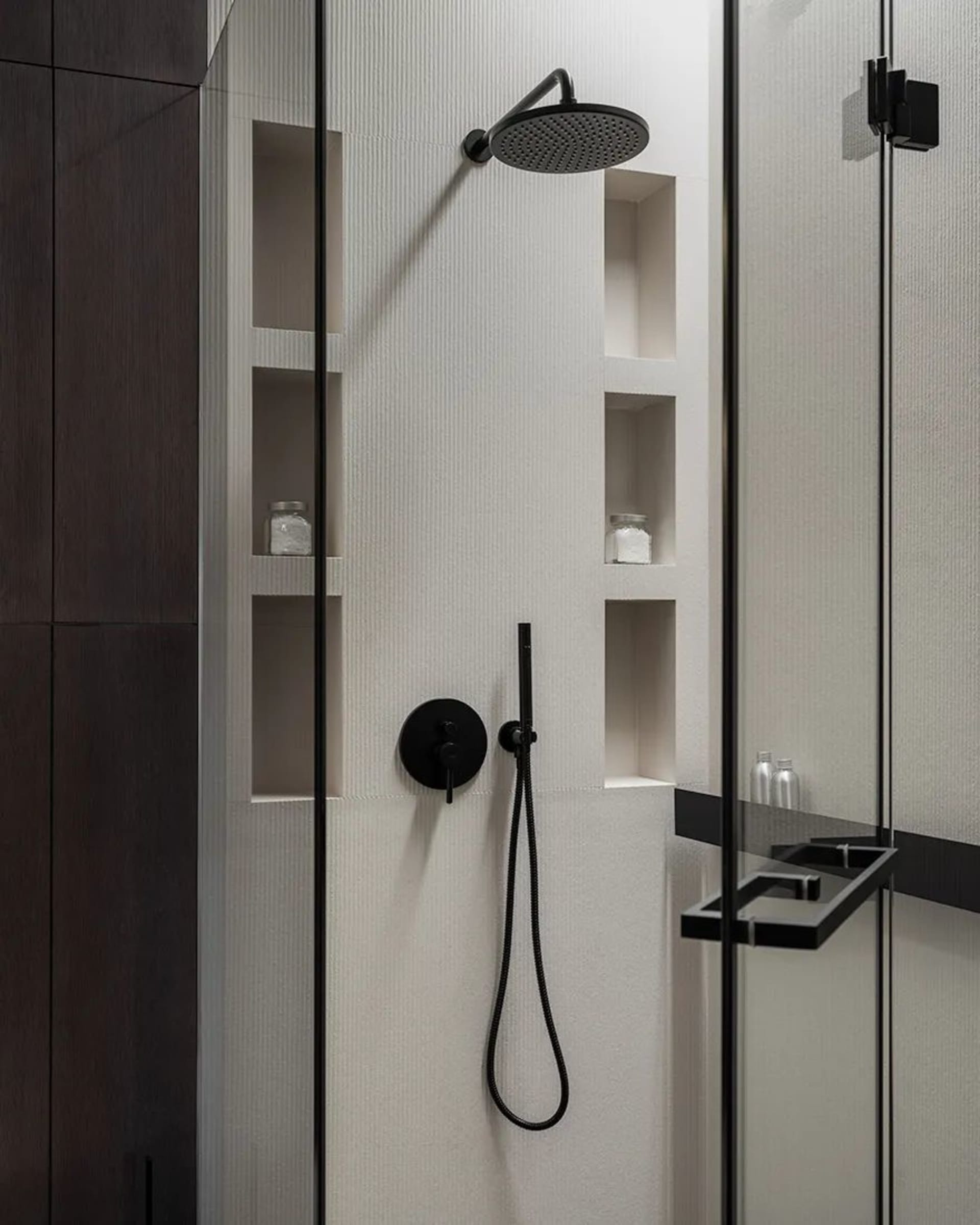 A modern minimalist bathroom featuring a textured white tiled wall and a glass partition. The bathroom includes a shower area
