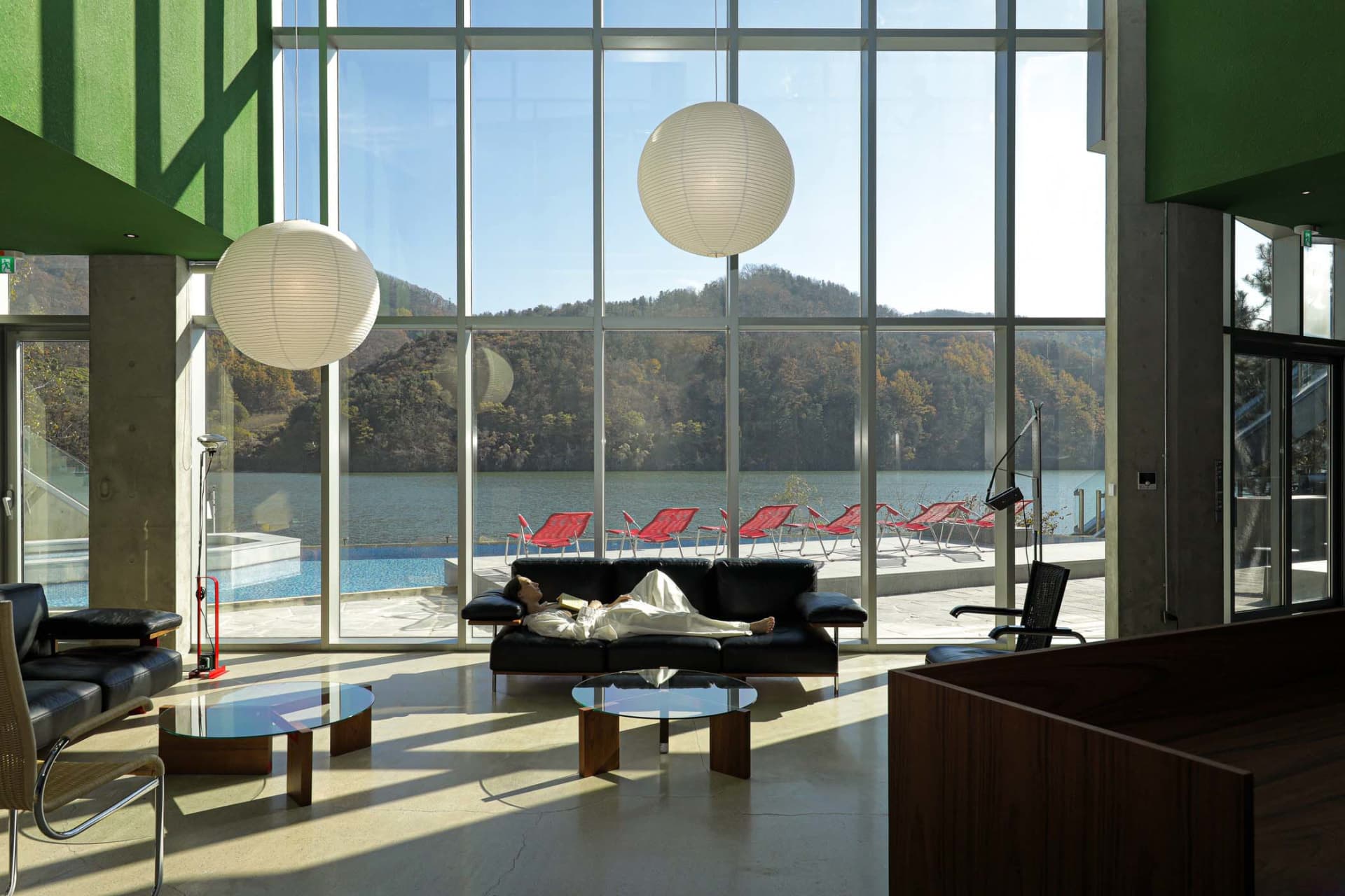 A modern hotel lobby features a glass wall overlooking a serene mountain lake, accompanied by black leather couches and red l