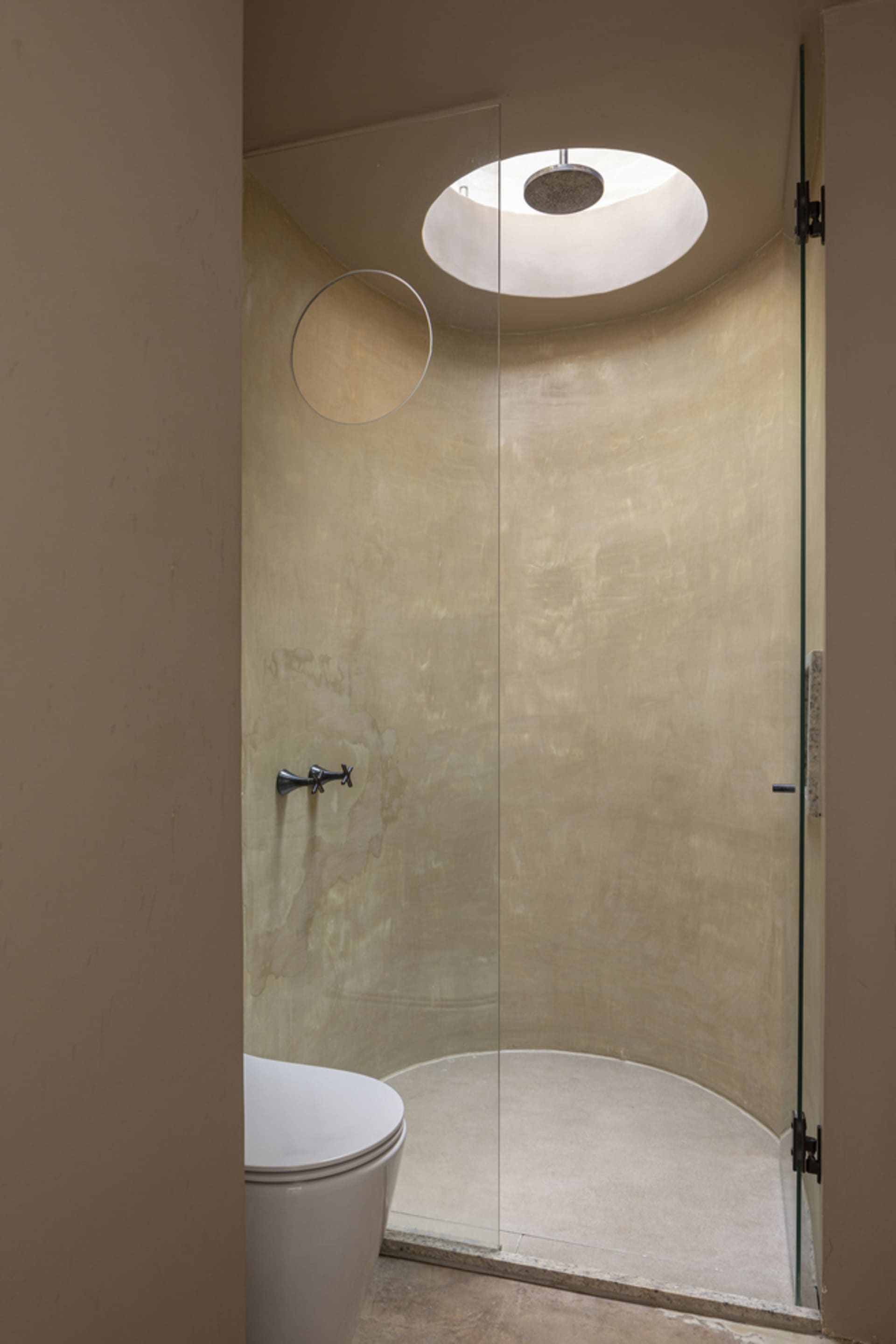 A minimalist bathroom featuring a concrete shower enclosure, circular skylight, and a white toilet.