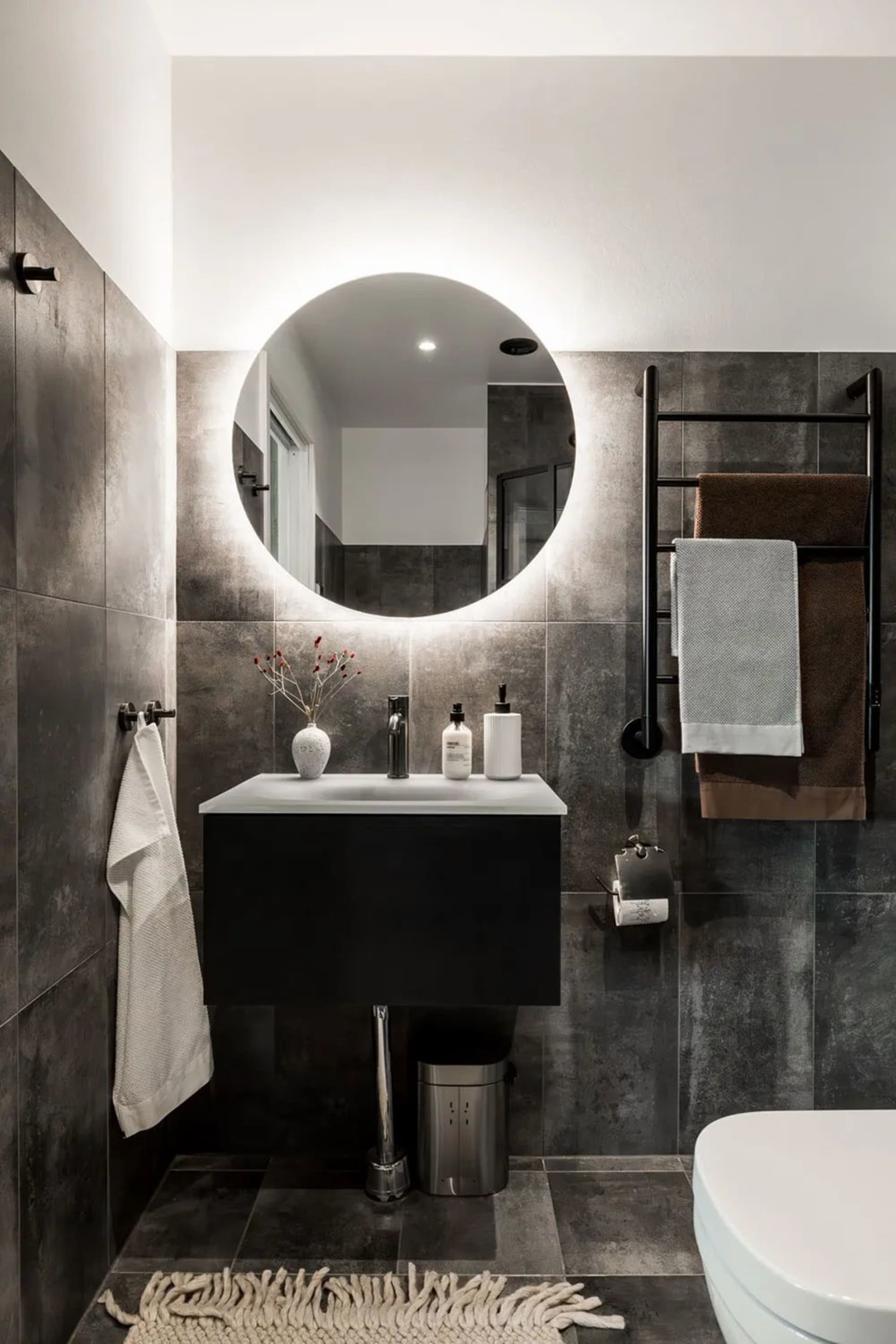 A modern minimalist bathroom with concrete walls and floor tiles, a floating vanity, and a round illuminated mirror.