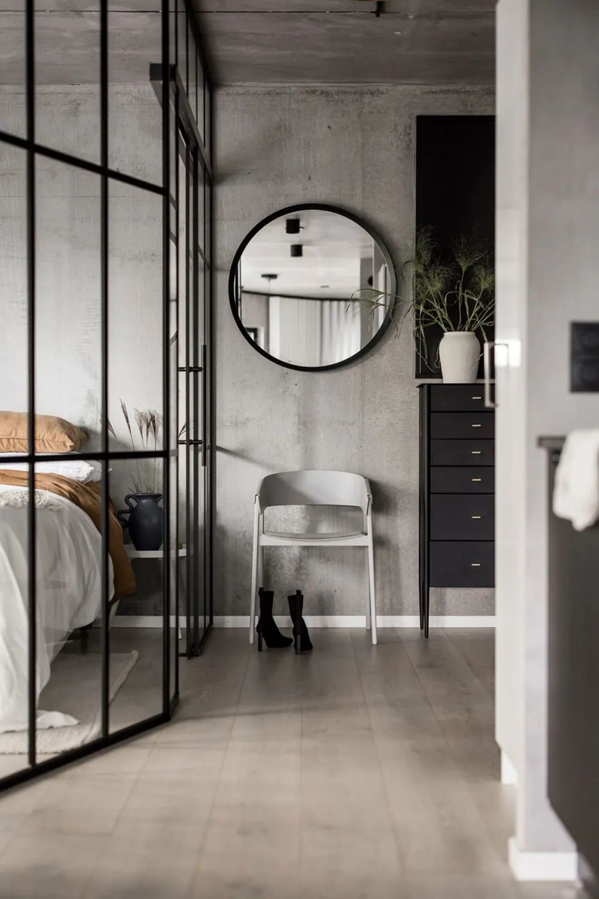 A minimalist bedroom with concrete walls, a glass partition, and a round mirror.