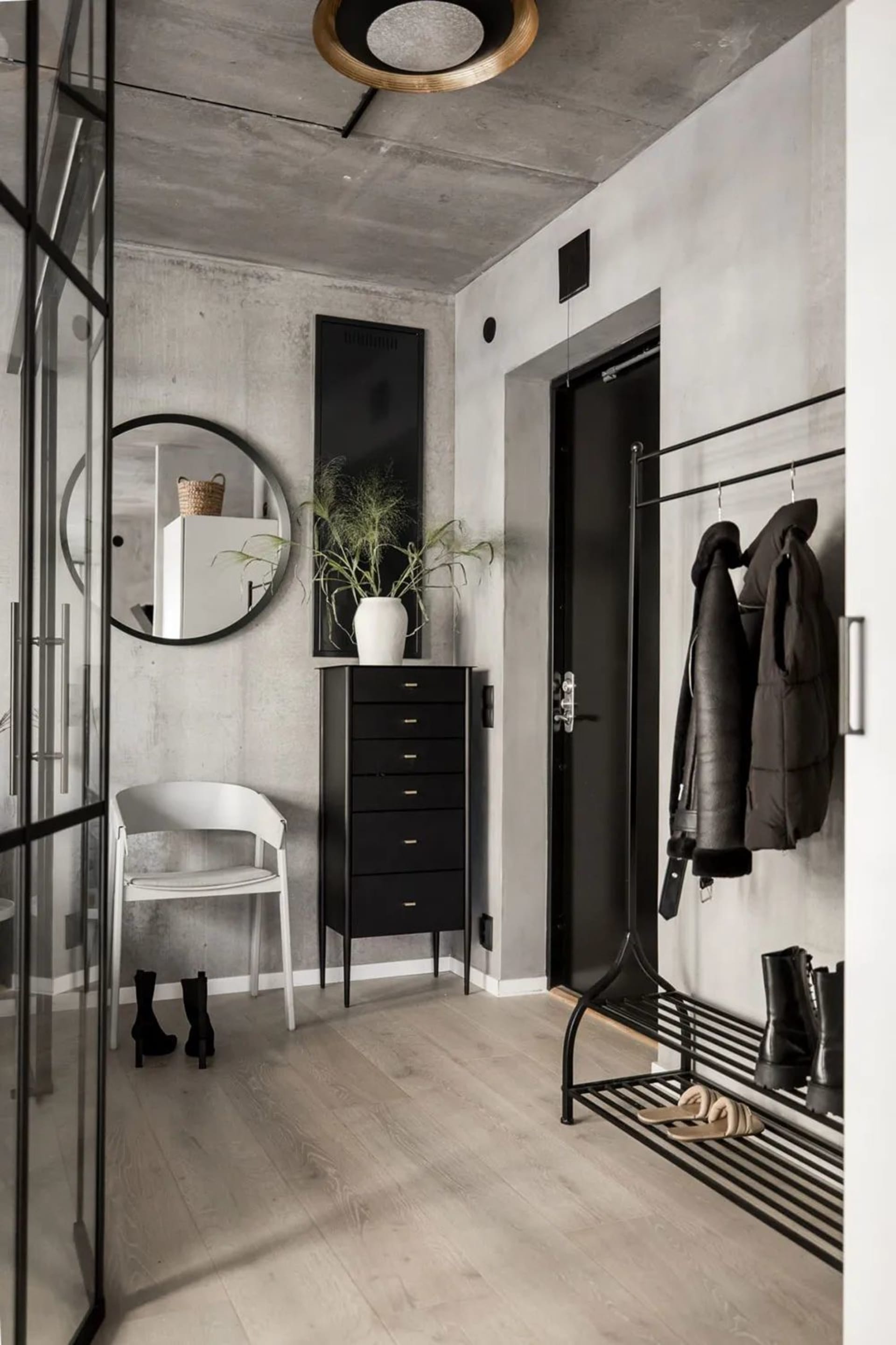 A modern, minimalist entryway featuring concrete, wooden, and metallic elements with a tall black cabinet, a white chair, and