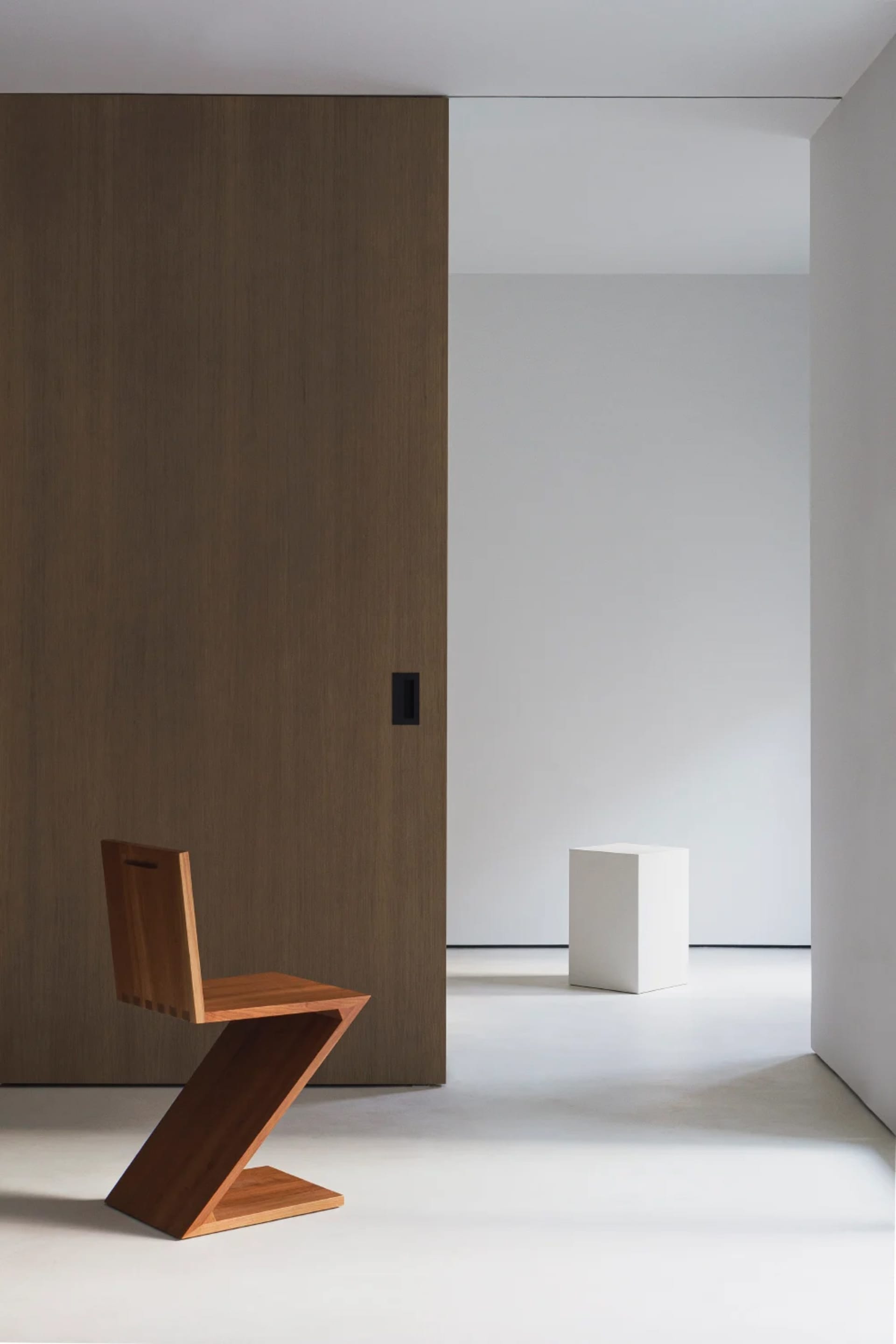 A minimalist space featuring a wooden zigzag chair and a white cube on a polished concrete floor.