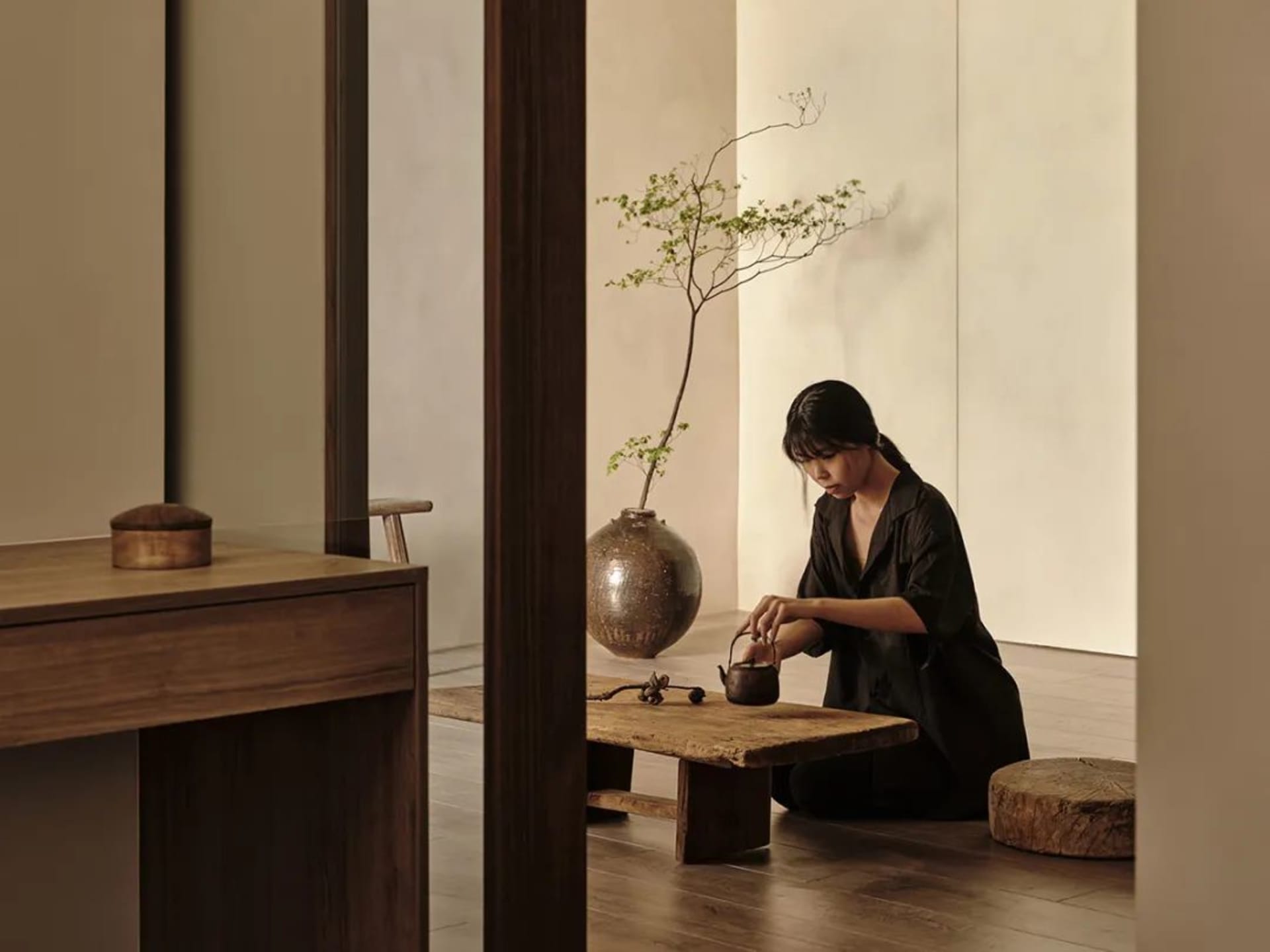 A person sitting cross-legged on the floor performing tea ceremony in a serene, minimalist room with wooden furniture and cer