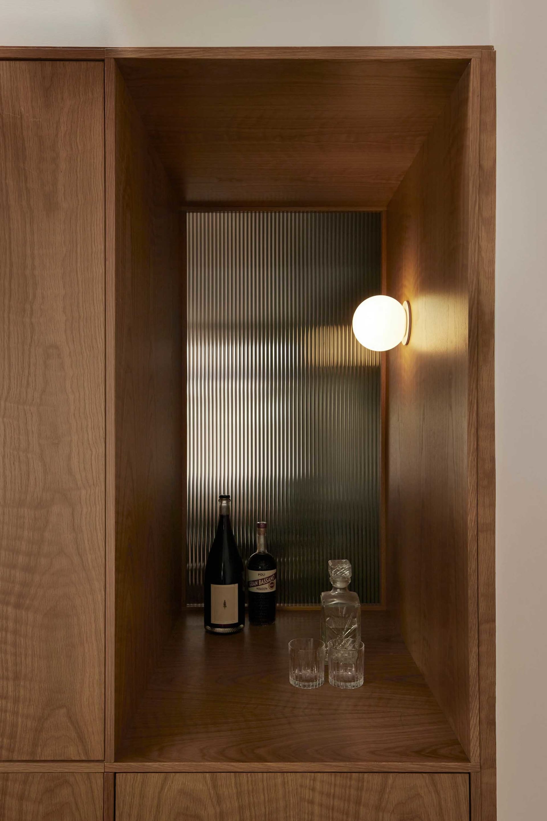 A modern bar shelf made of wood with a spherical wall-mounted light fixture, bottles of wine and liquor, and glasses.