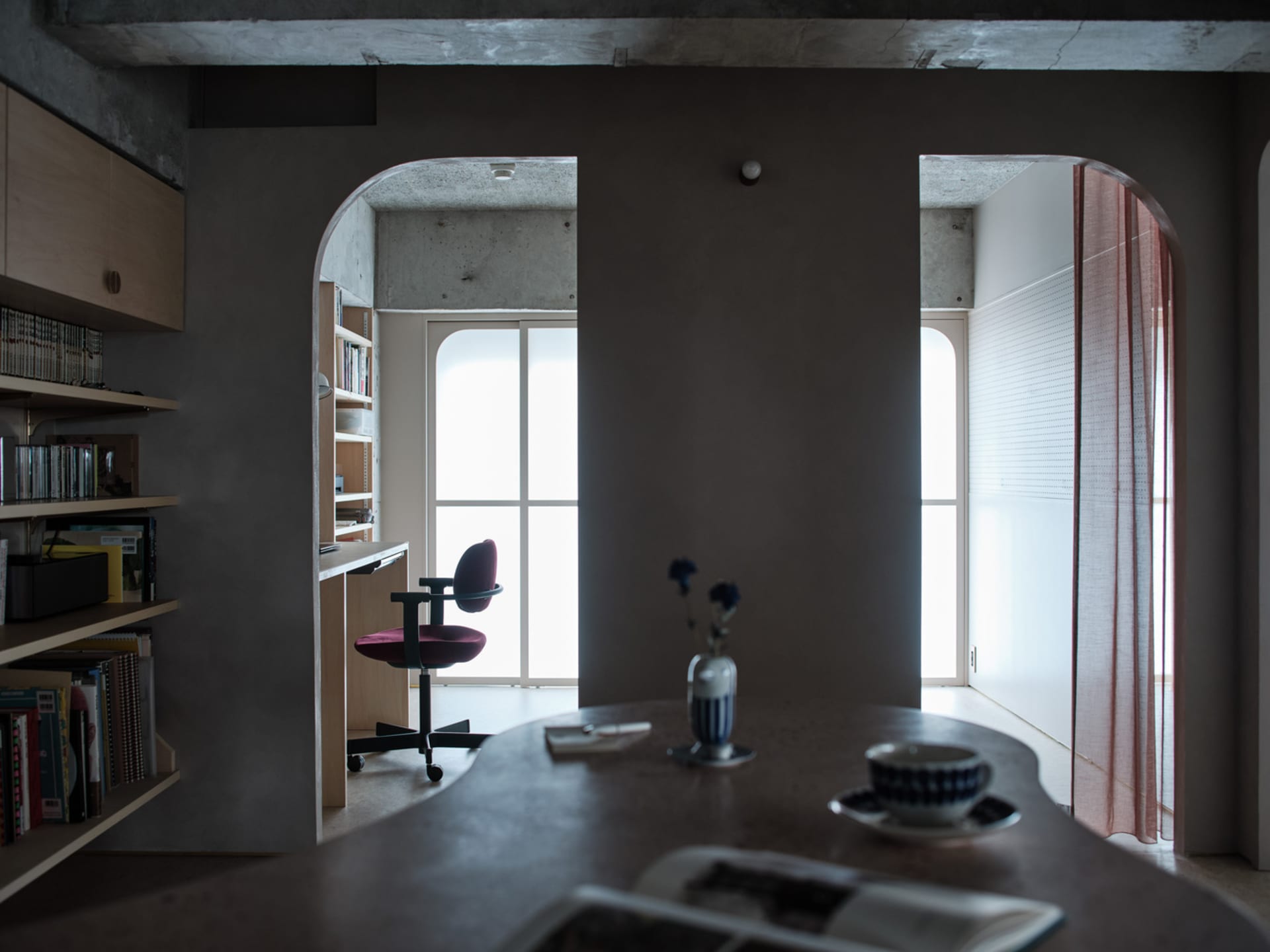 A modern minimalist home office features a concrete wall with an arched doorway leading to a wooden desk.