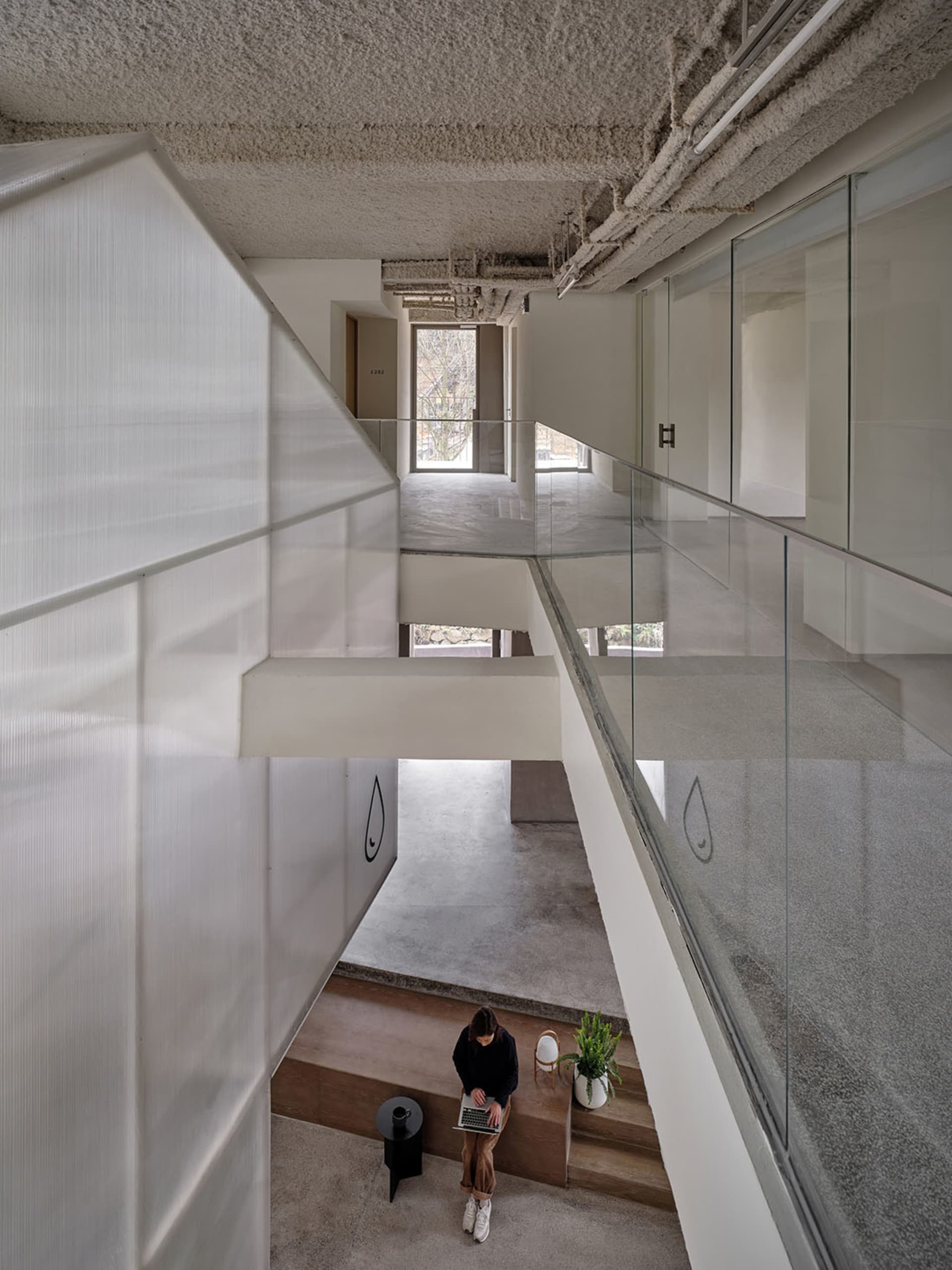 A modern office space with concrete floors and walls, featuring a glass partition and a person working on a laptop.