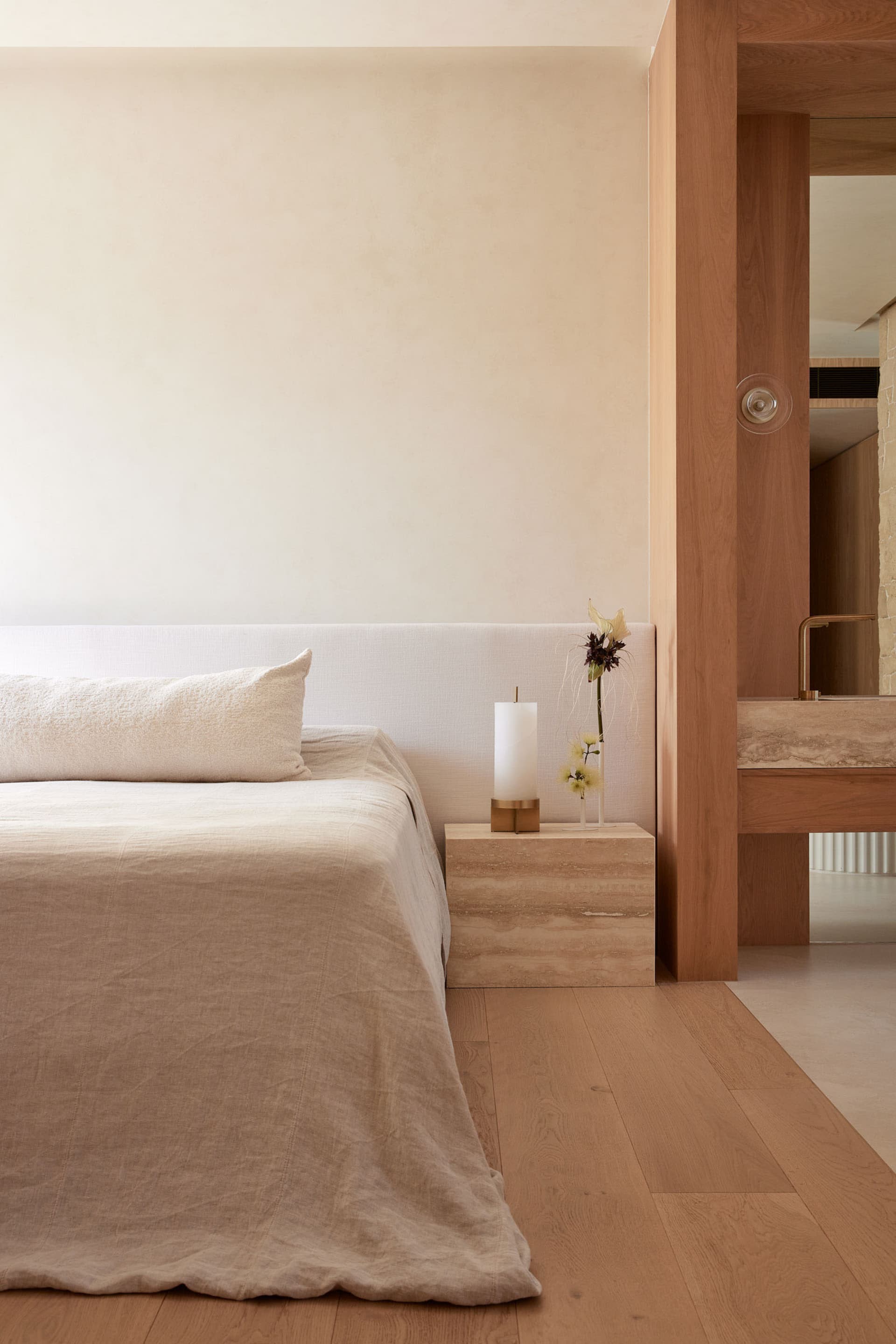 Minimalist bedroom with a wooden floor, beige bedding, and a wooden nightstand with a candle and a vase of flowers.