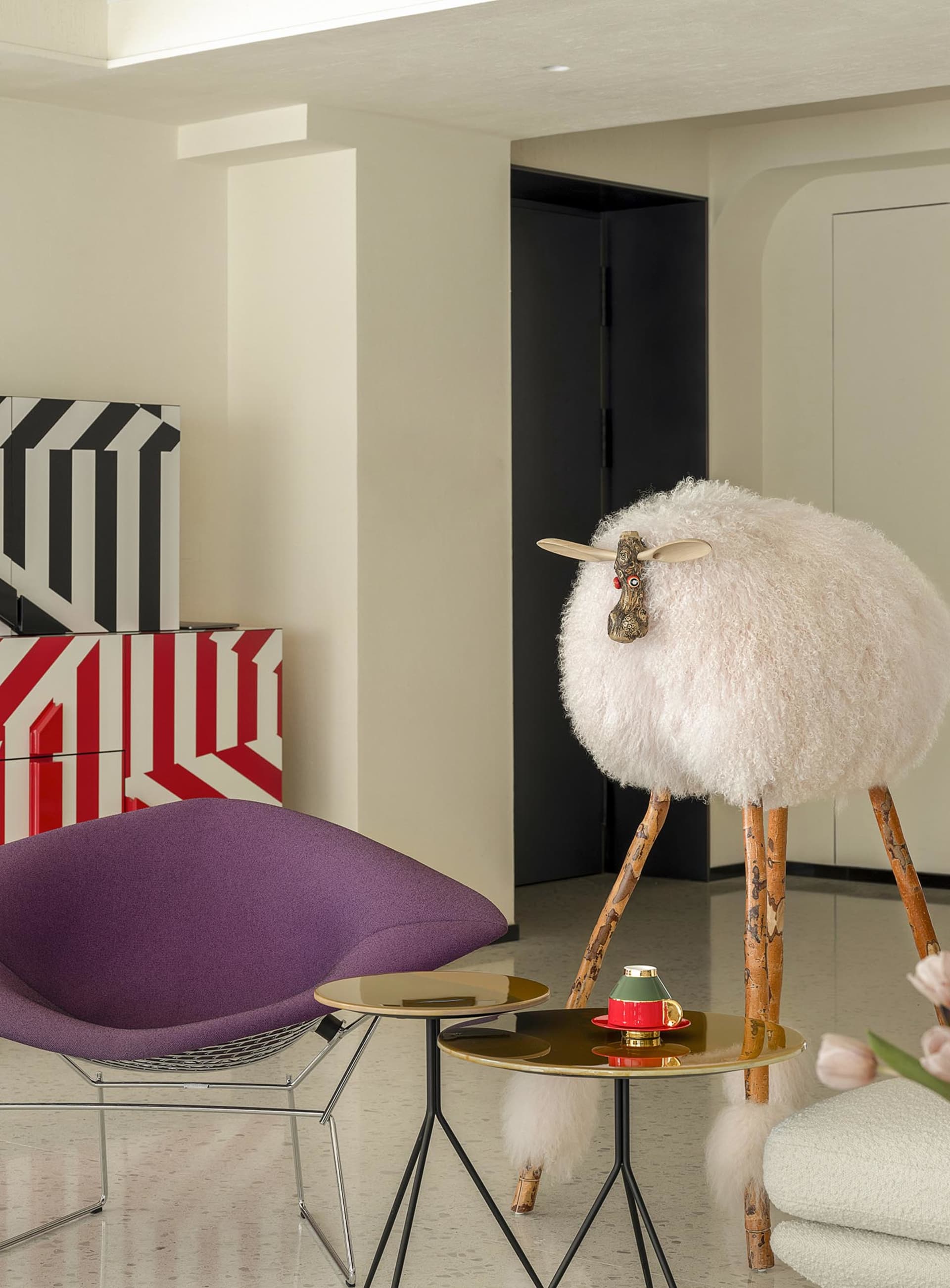 A modern living room with a purple fabric chair, a sheep sculpture, and a side table with a coffee cup and vase of pink tulip