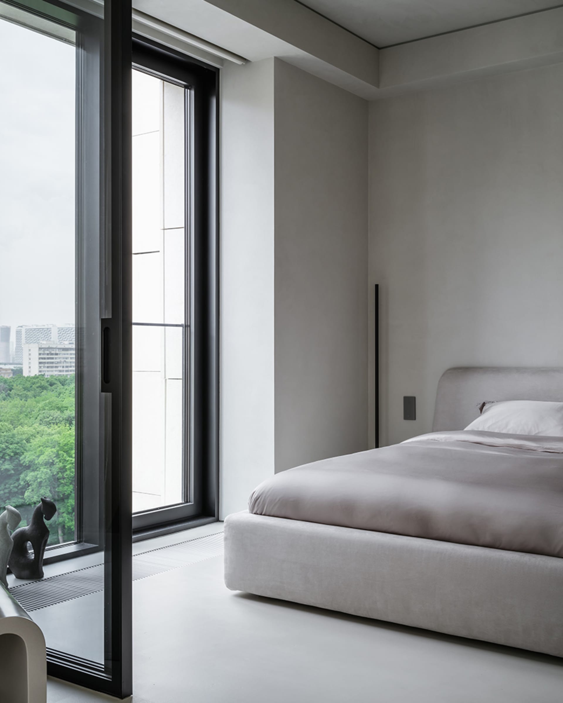 Modern minimalist bedroom featuring a grey upholstered bed with a fabric headboard, large window with urban view, and sleek d
