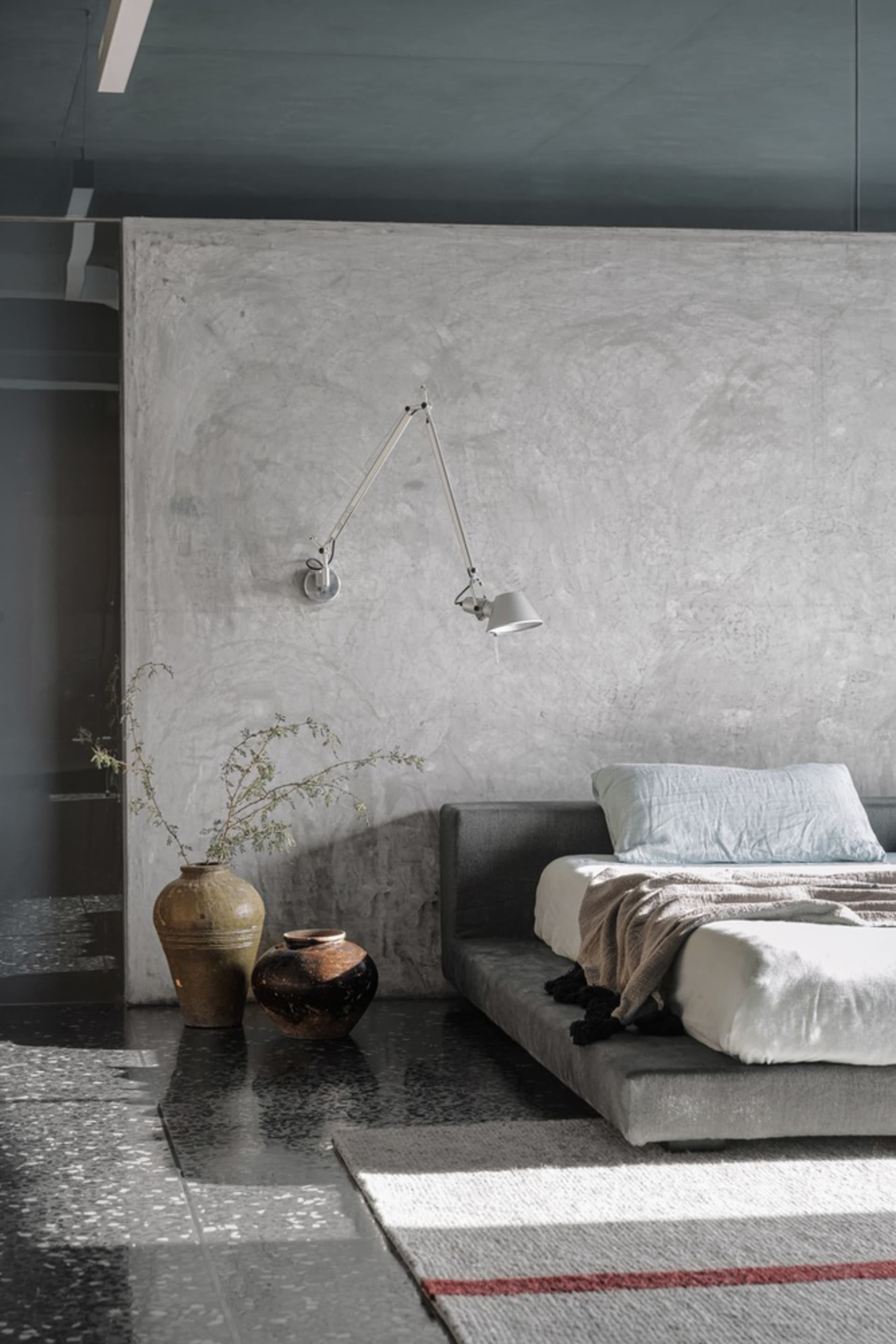 A minimalist bedroom with a concrete wall, a bed, a wall lamp, and decorative vases.