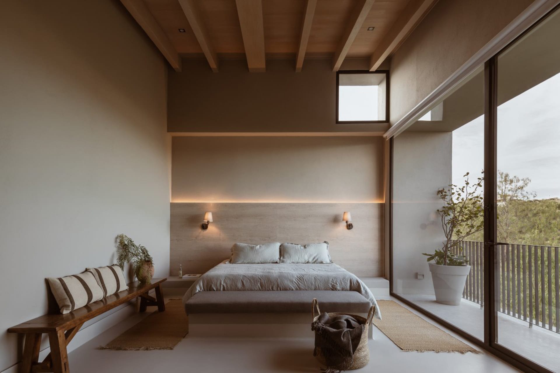 Modern bedroom featuring a wooden bed and wall-mounted lamps, with a plant near the window and a bench with striped pillows.