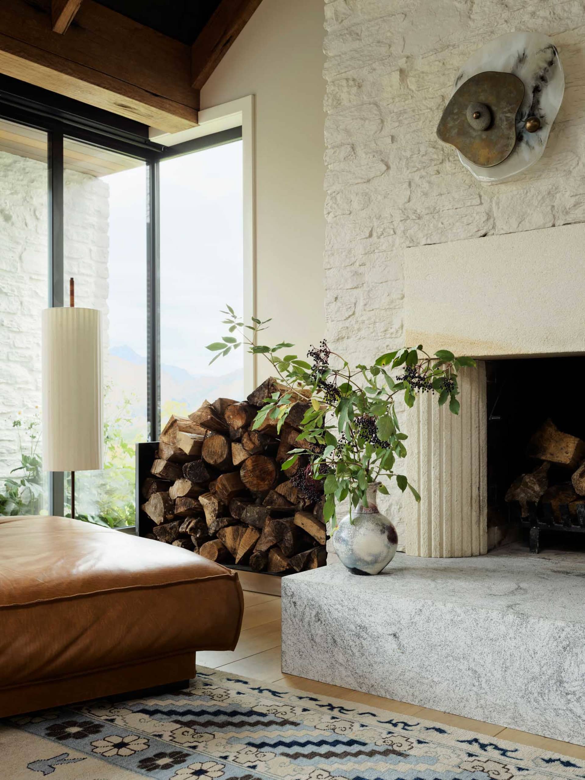 A stone fireplace in a living room with wood logs and a vase of greenery on the mantle.