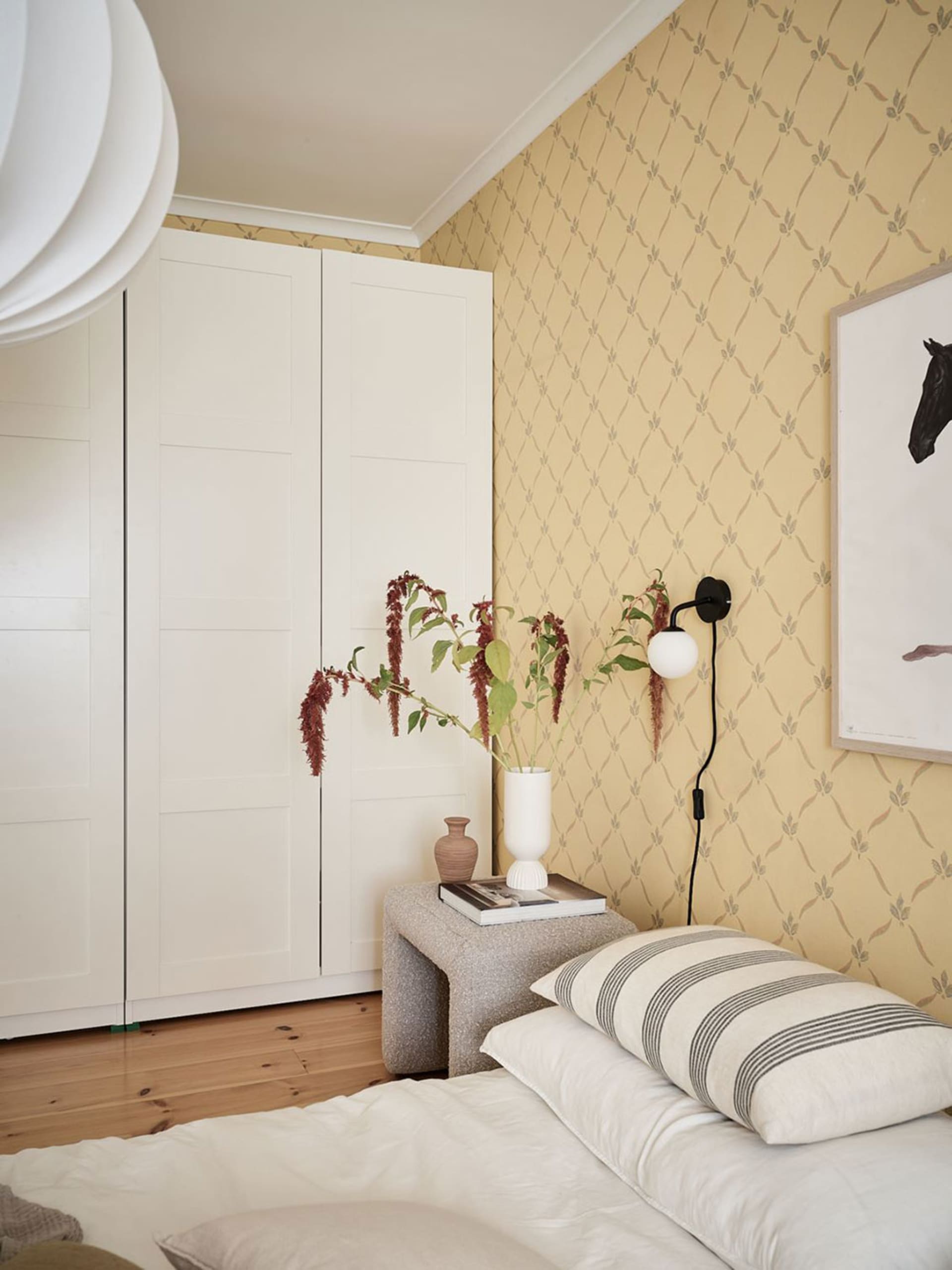 Minimalist bedroom with neutral tones featuring a bed, pillows, and a small bench with a white vase and flowers.