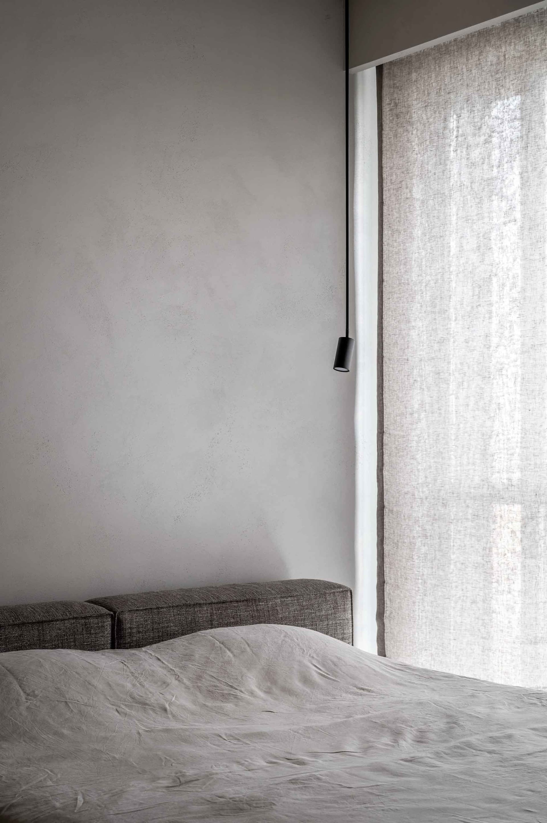 A modern grey bedroom featuring a bed draped in light grey fabric, minimalistic design, and natural light filtering through s