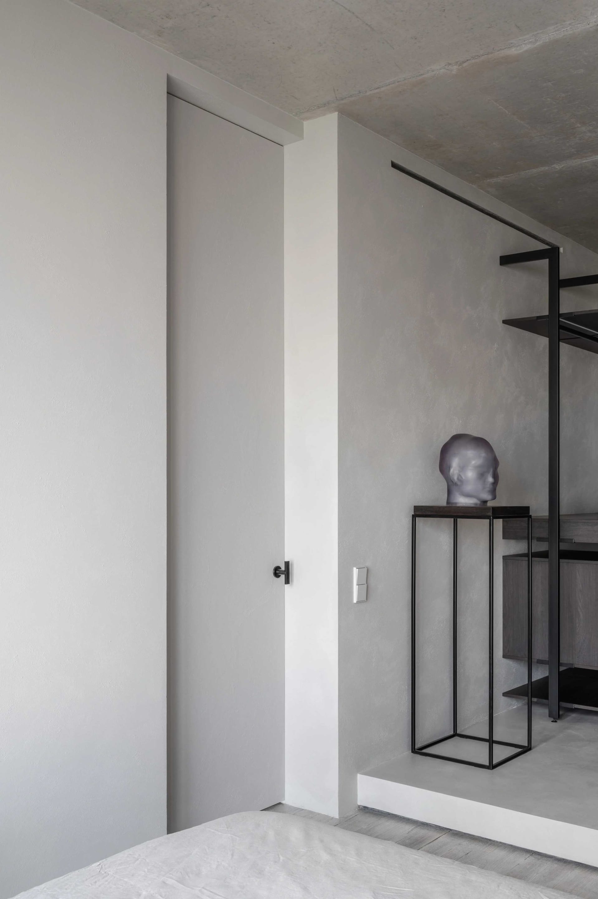 A minimalist bedroom featuring a concrete wall and floor, with a bed and a sculpture on a pedestal.