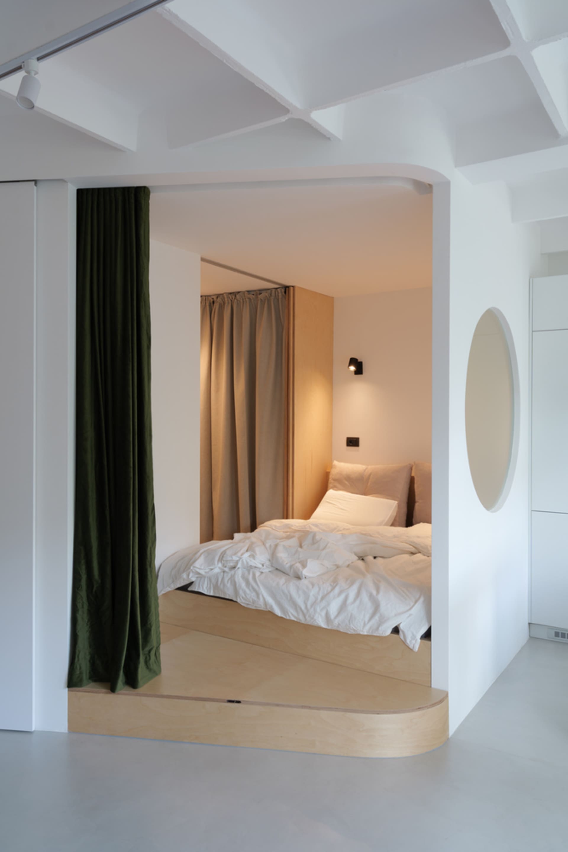 A modern minimalist bedroom with a bed platform embedded in the floor, surrounded by white walls, wood panels, and layered cu