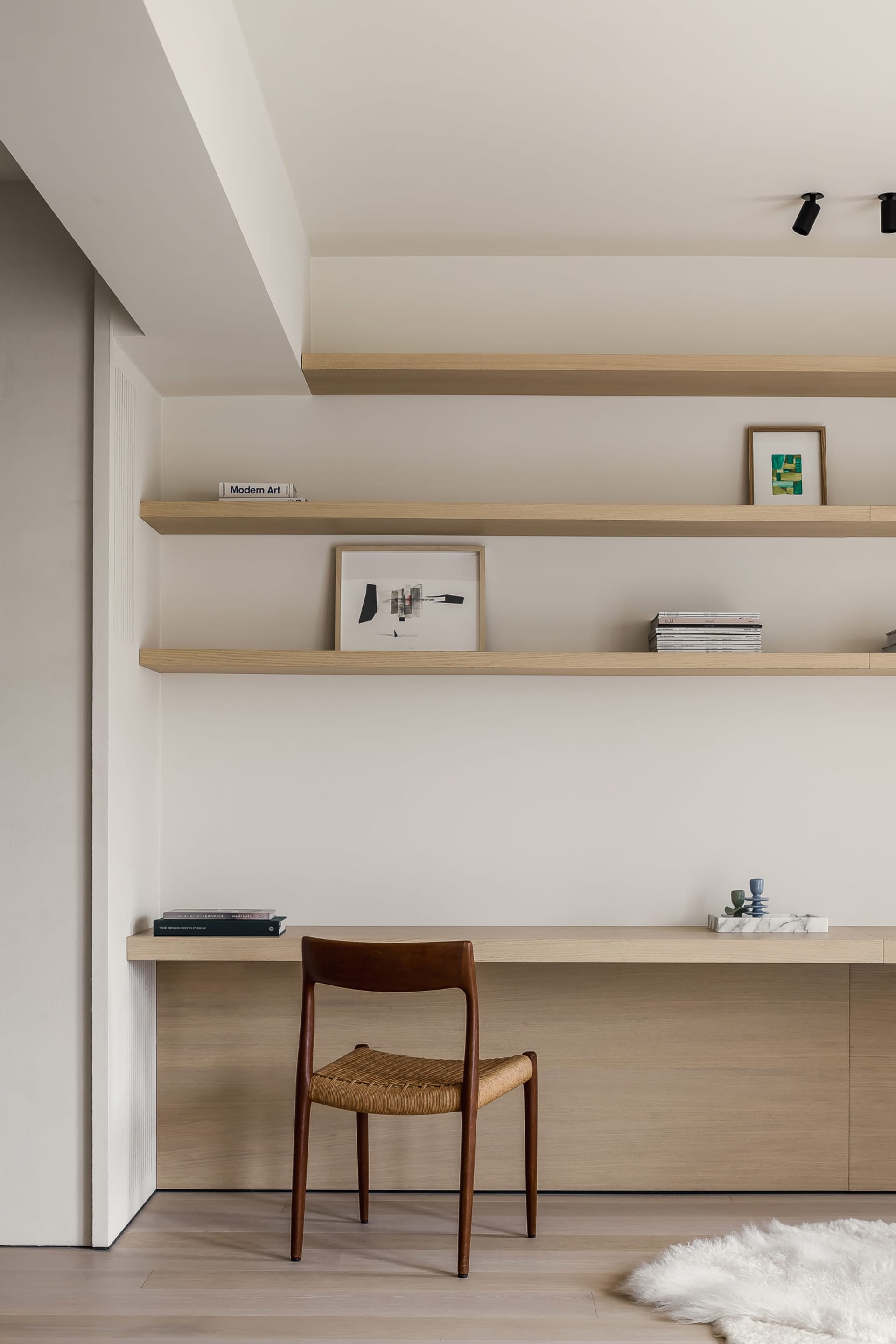 A modern home office featuring floating shelves with books and art, a wooden chair, and minimalist design.