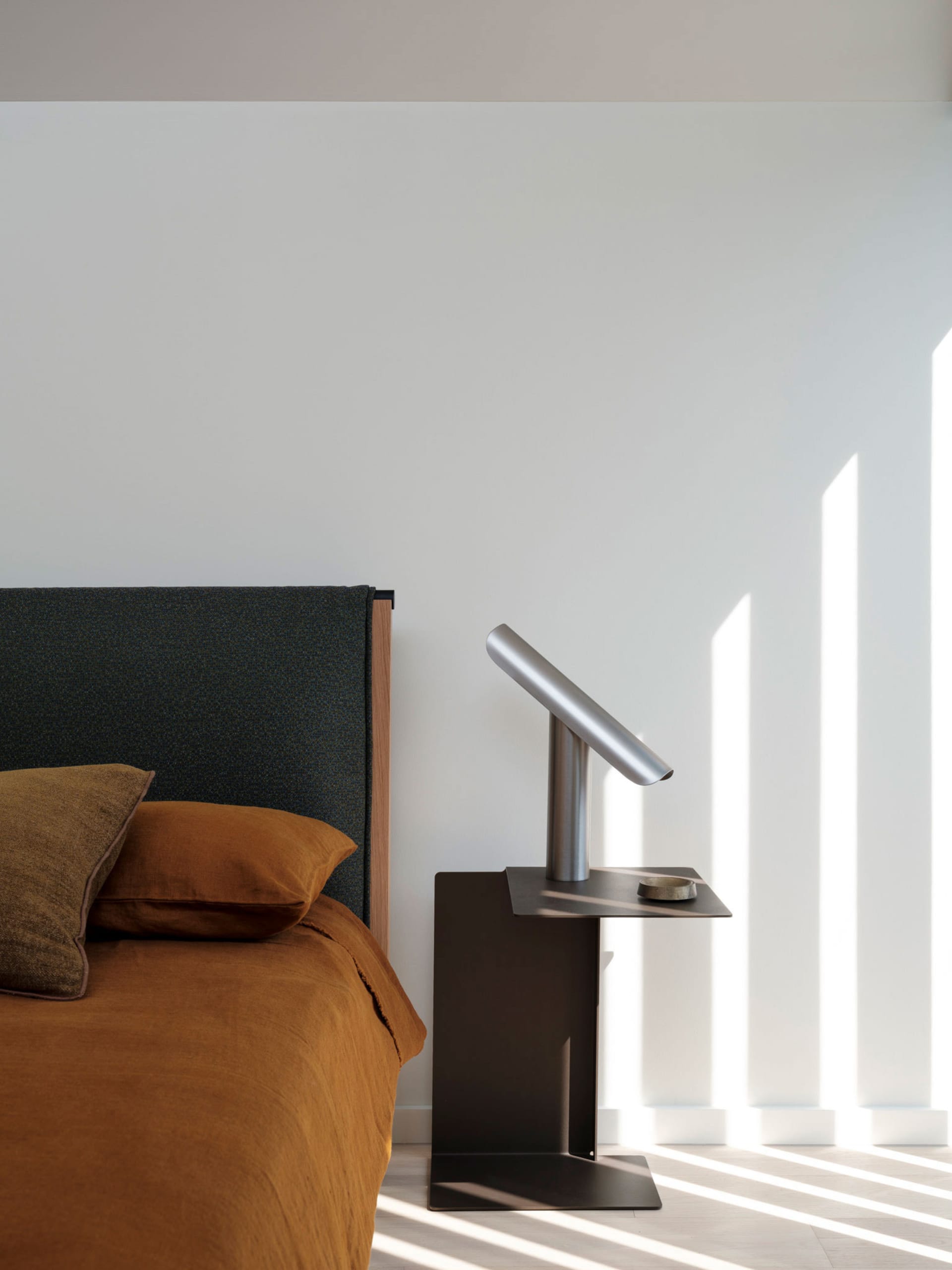 A minimalistic bedroom with a grey upholstered bed headboard and orange bedding, featuring a sleek, metallic bedside lamp wit