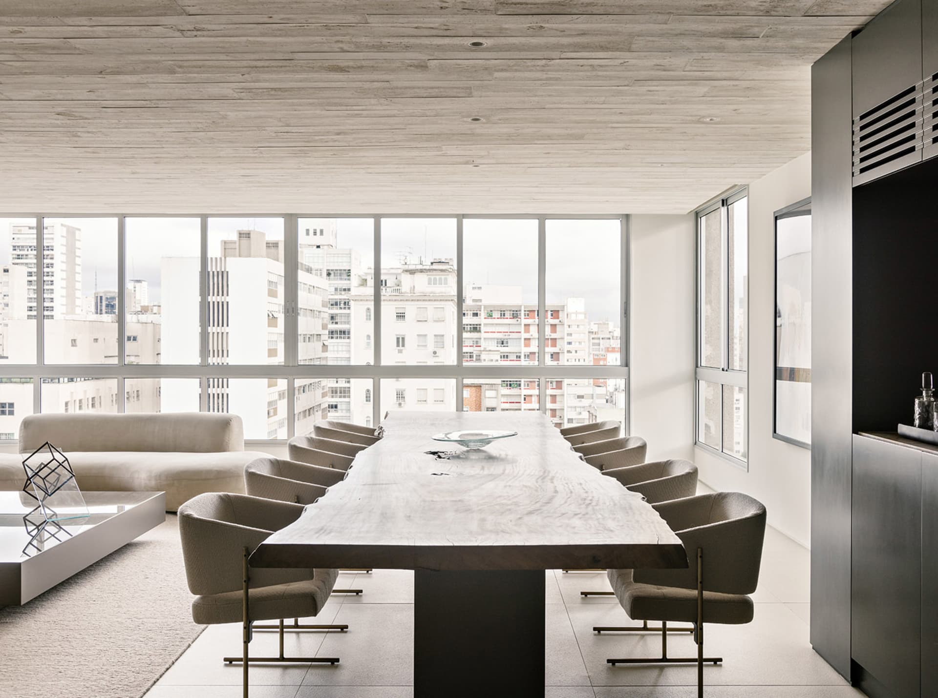 A modern conference room featuring a long wooden table surrounded by upholstered chairs and a large window with a cityscape v