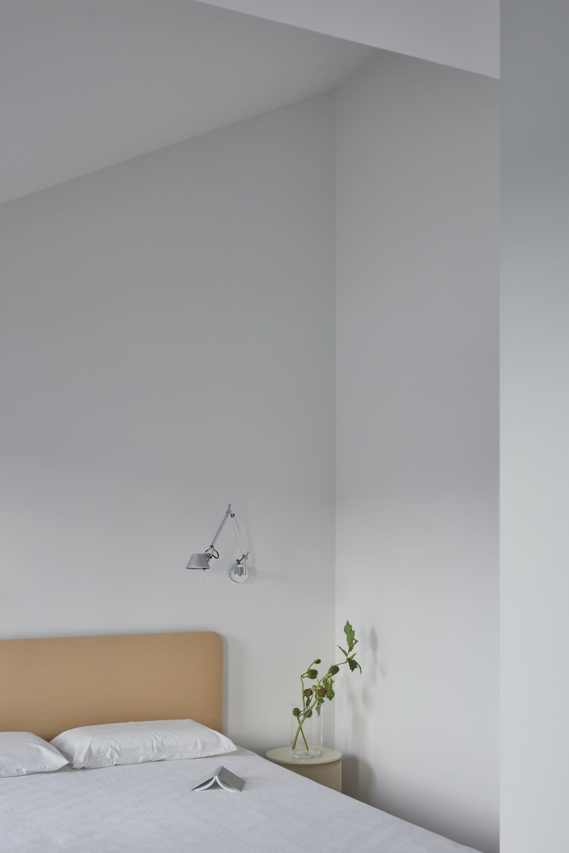 A modern minimalist bedroom featuring a bed with a fabric headboard and white bedding, placed in front of a white wall with a