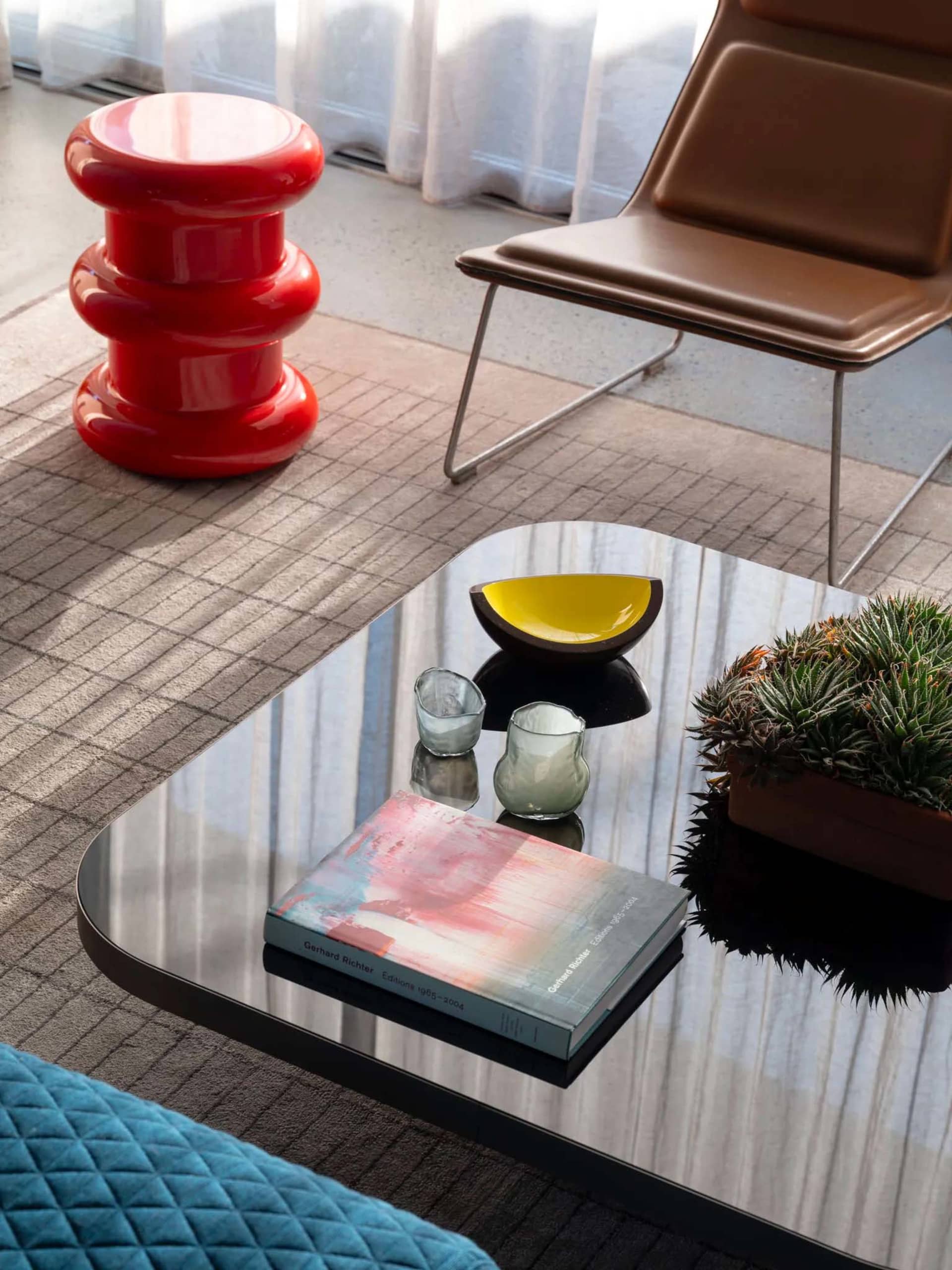 Modern living room featuring a glass coffee table with a red sculptural stool, yellow and green glass bowls, and a potted pla