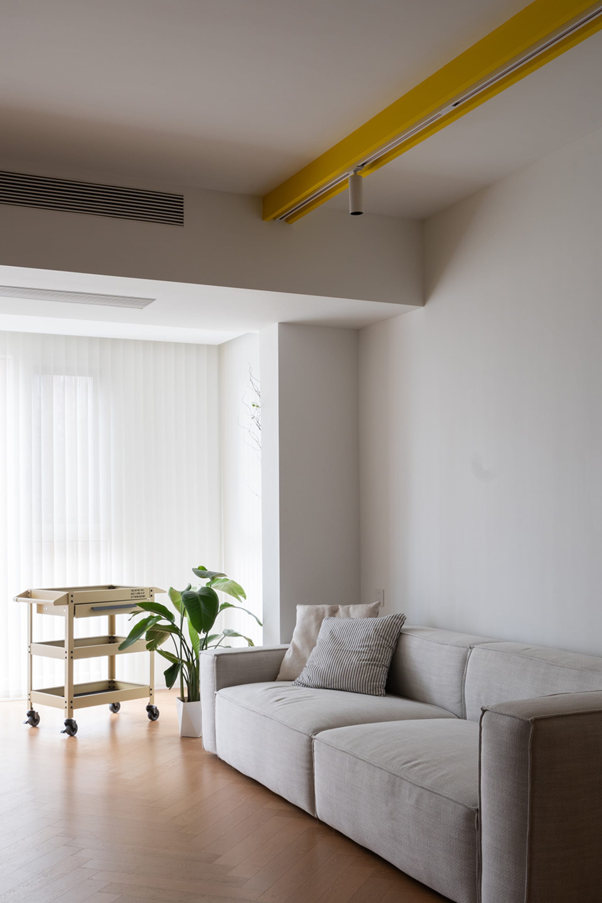 Modern minimalist living room featuring a fabric couch with a yellow track light overhead and a rolling cart with a plant.