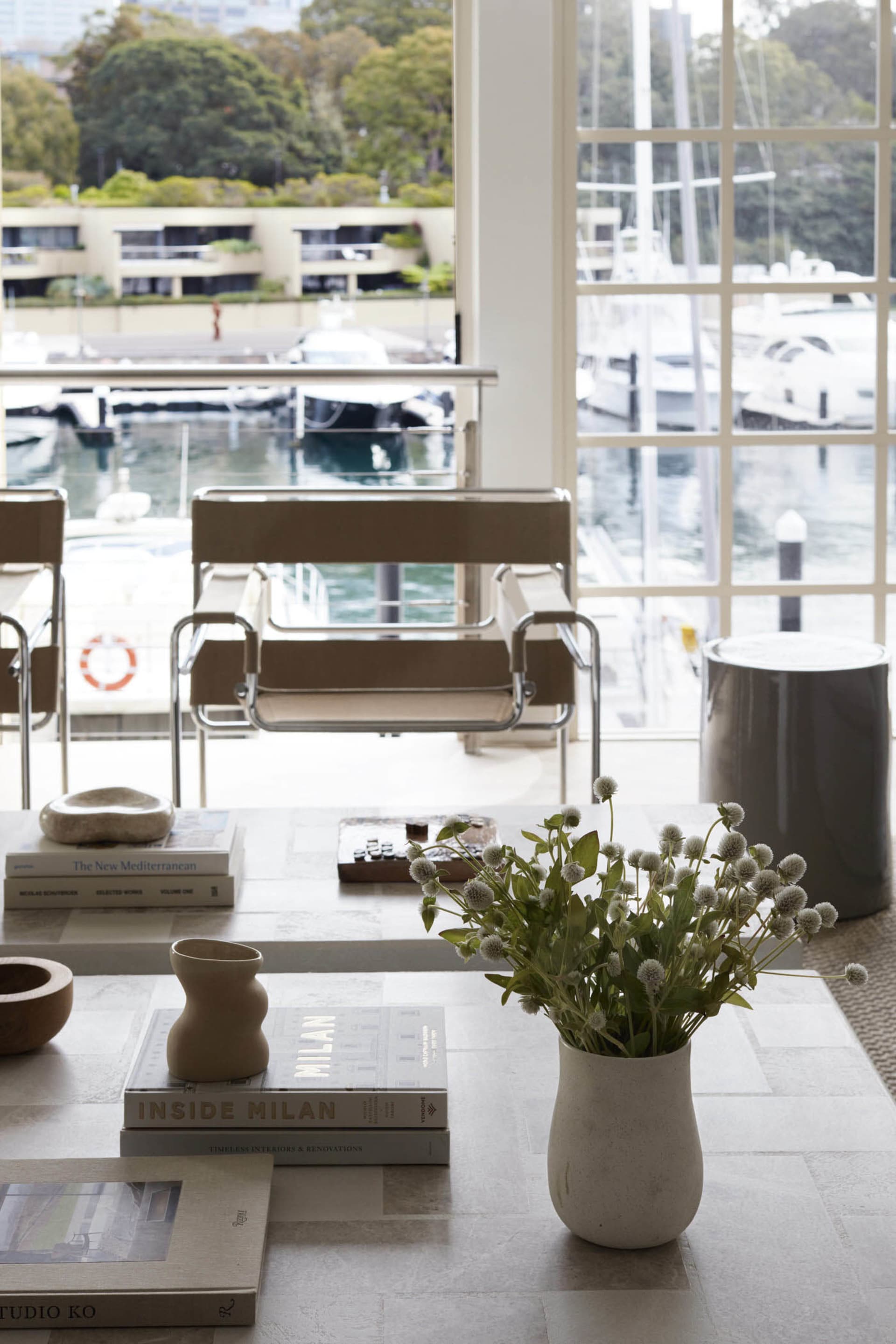 A modern living room featuring wooden furniture, books, and a vase of flowers with white blooms on a table by a large window