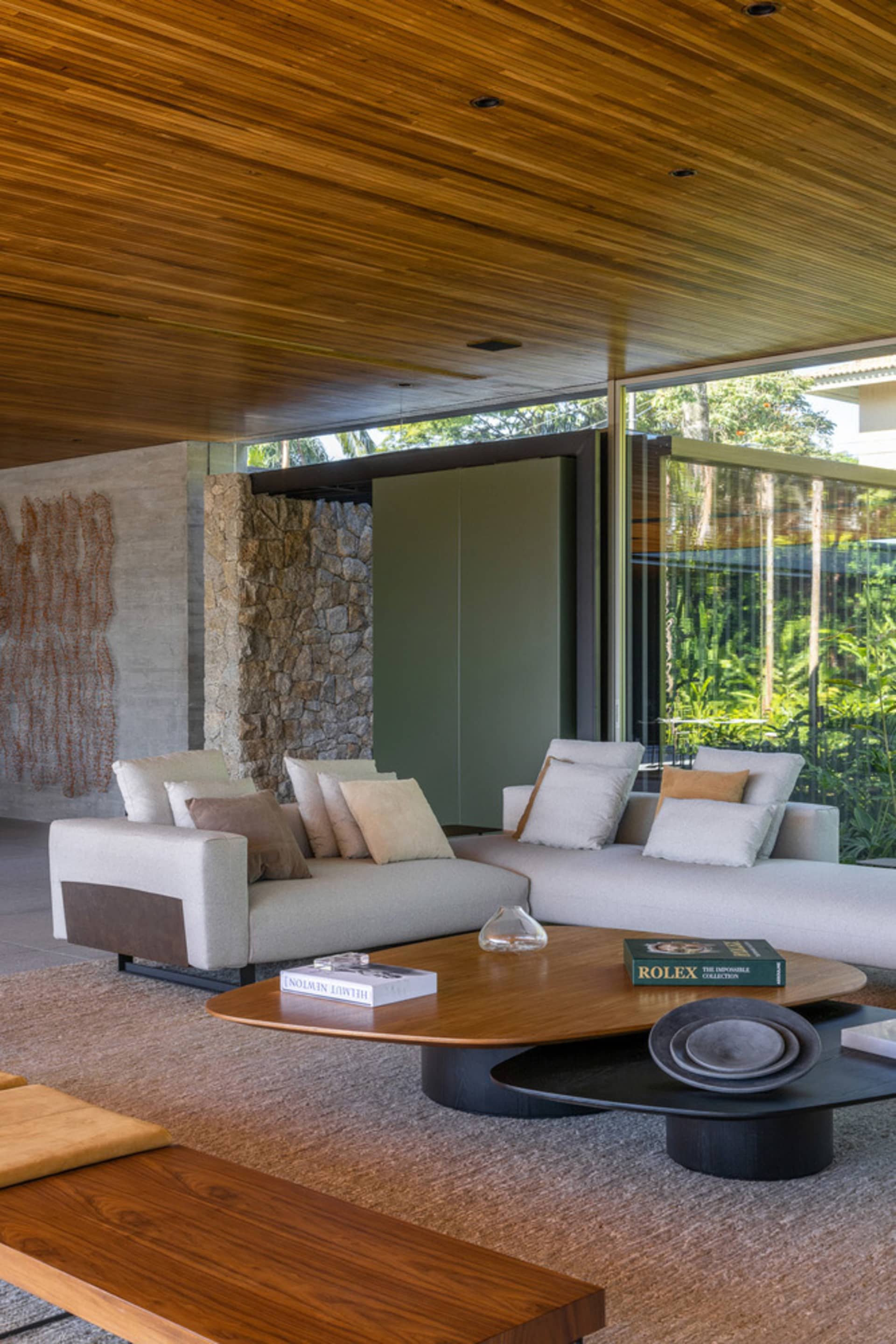 A modern industrial-style living room with wooden ceiling, sofa, and tables, featuring a stone fireplace and large windows.