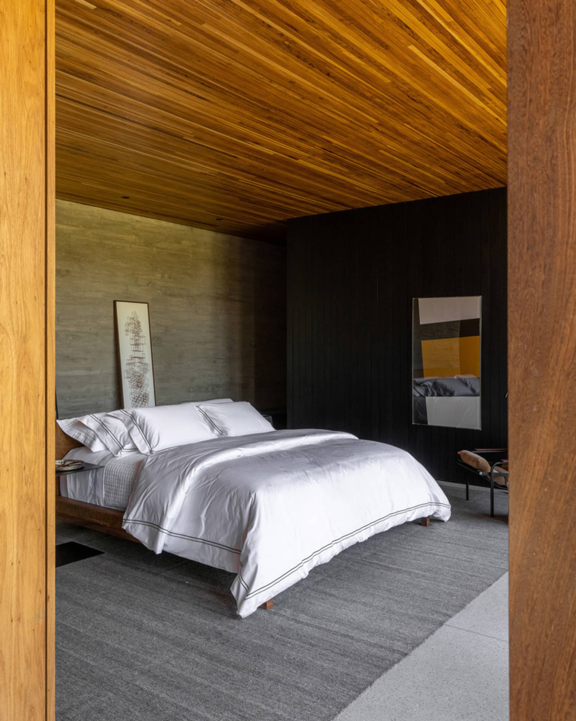 A minimalist bedroom with a white bed, grey carpet, and wooden ceiling.