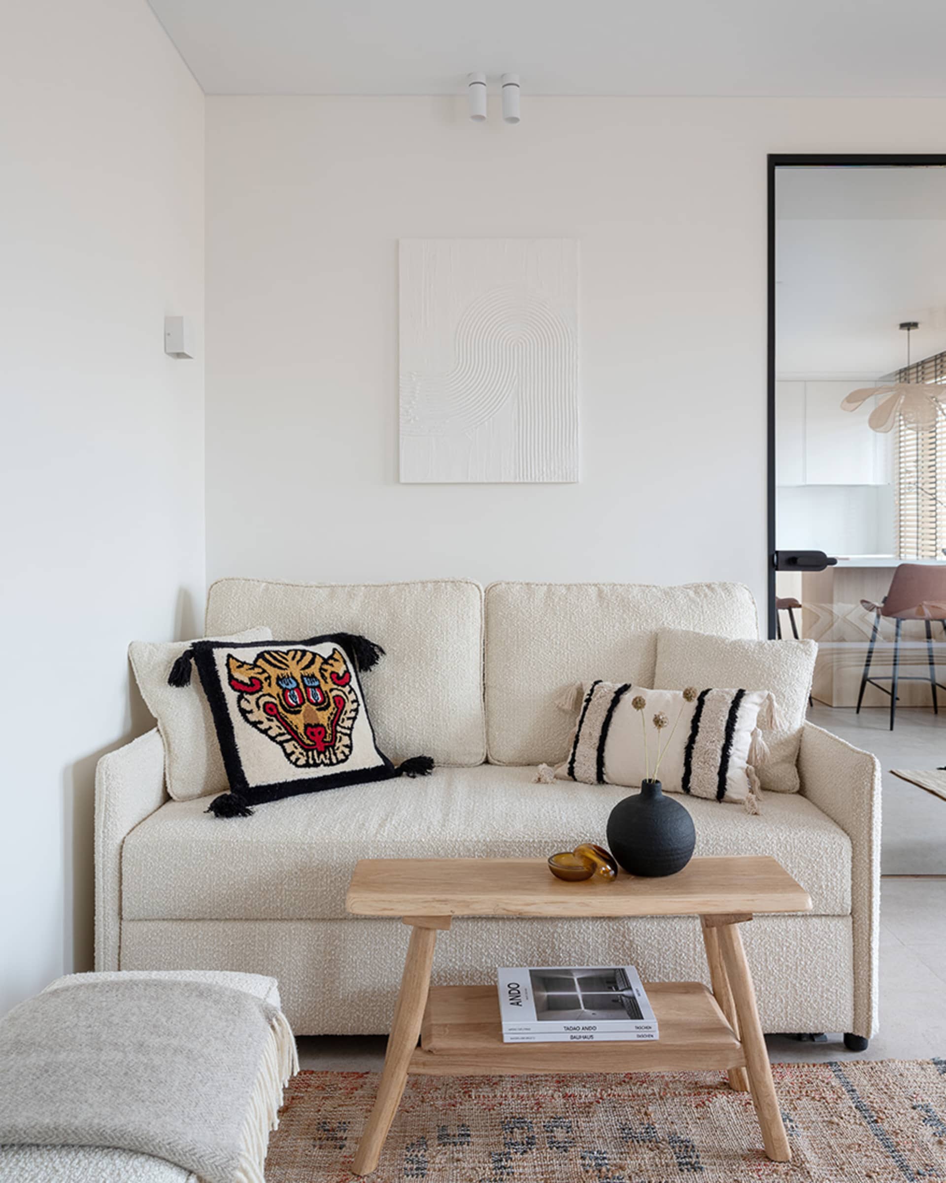 A minimalist living room featuring a neutral-toned sofa with decorative pillows, a wooden coffee table, and a rug, with a sim