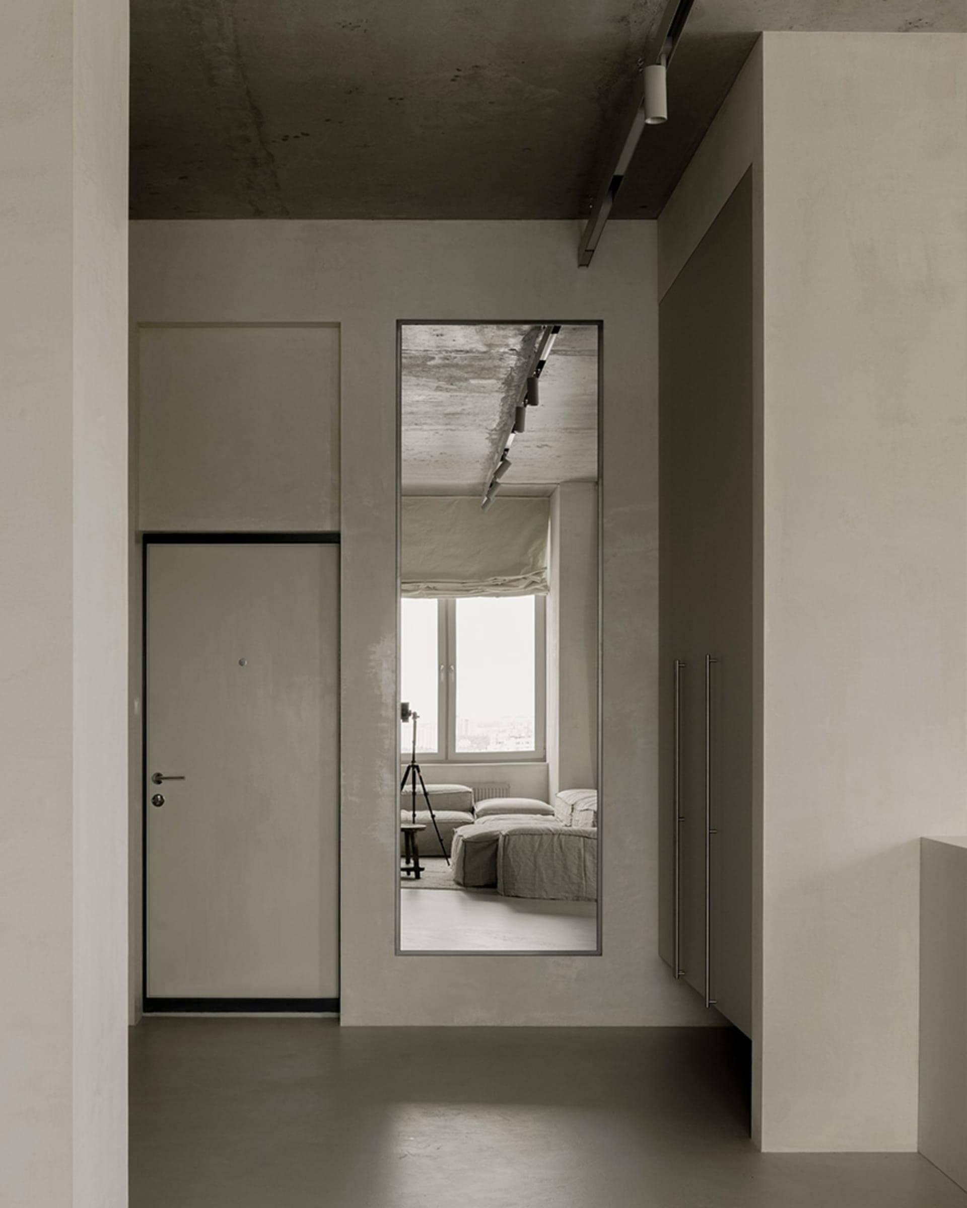 A minimalist living room with concrete walls and floor, featuring a large mirror reflecting the interior.