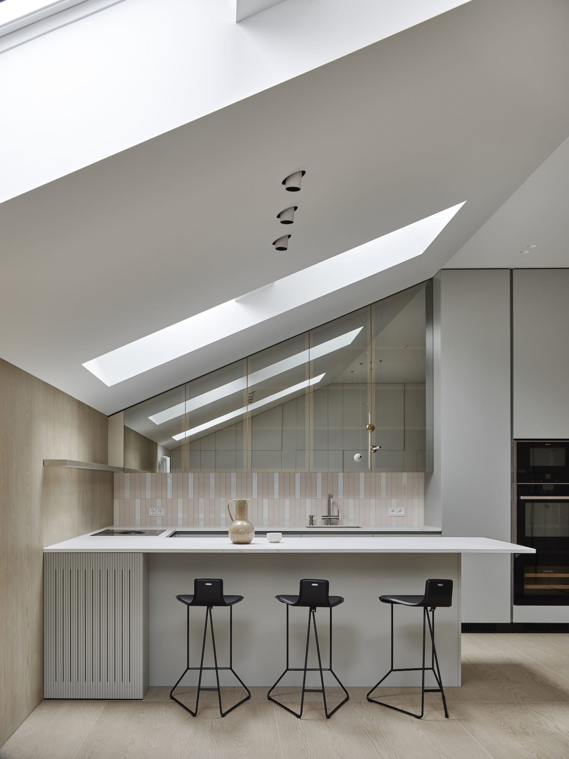 A modern residential kitchen with white and light-colored finishes, featuring skylights, a minimalist island, and black stool