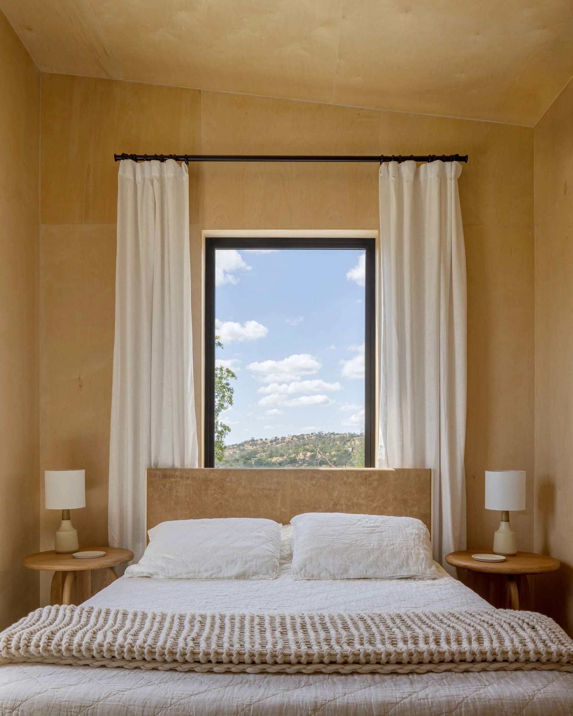 A minimalist bedroom with white curtains, a bed, and two side tables with lamps.