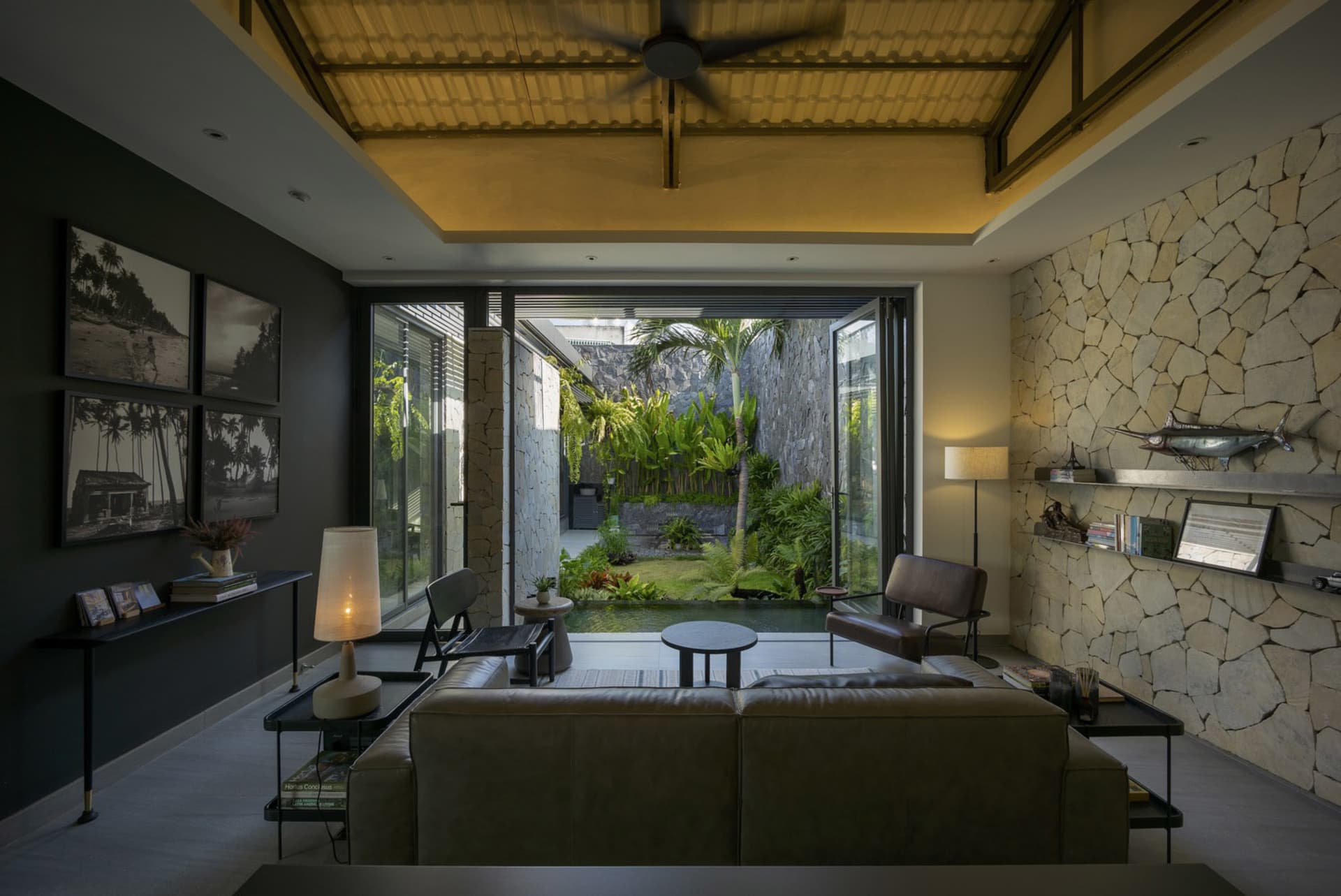 Modern living room featuring a concrete and leather sofa, stone wall, and tropical outdoor view.