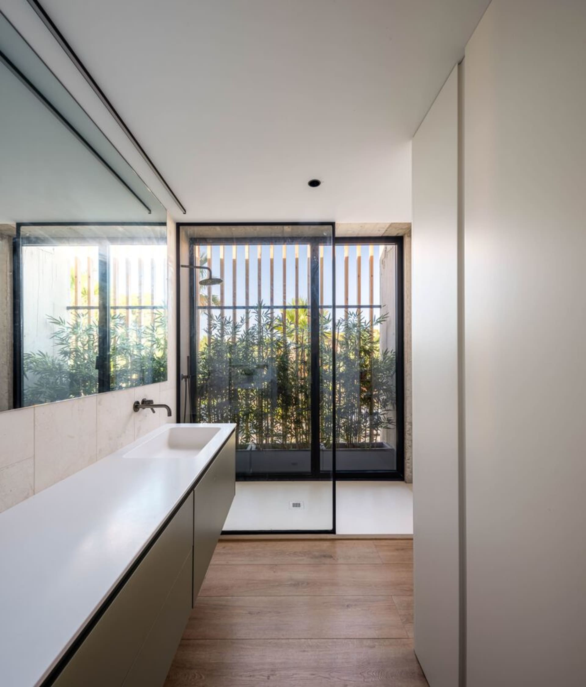 A modern bathroom with glass walls and wooden flooring, featuring a large window with a view of greenery.