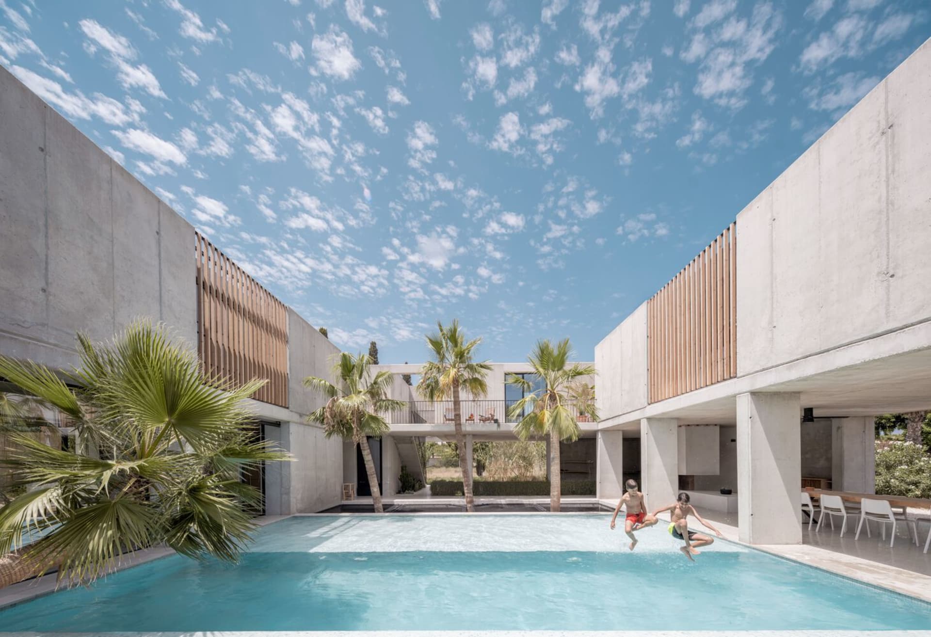 Two people jumping into a swimming pool surrounded by concrete walls with large windows and palm trees.