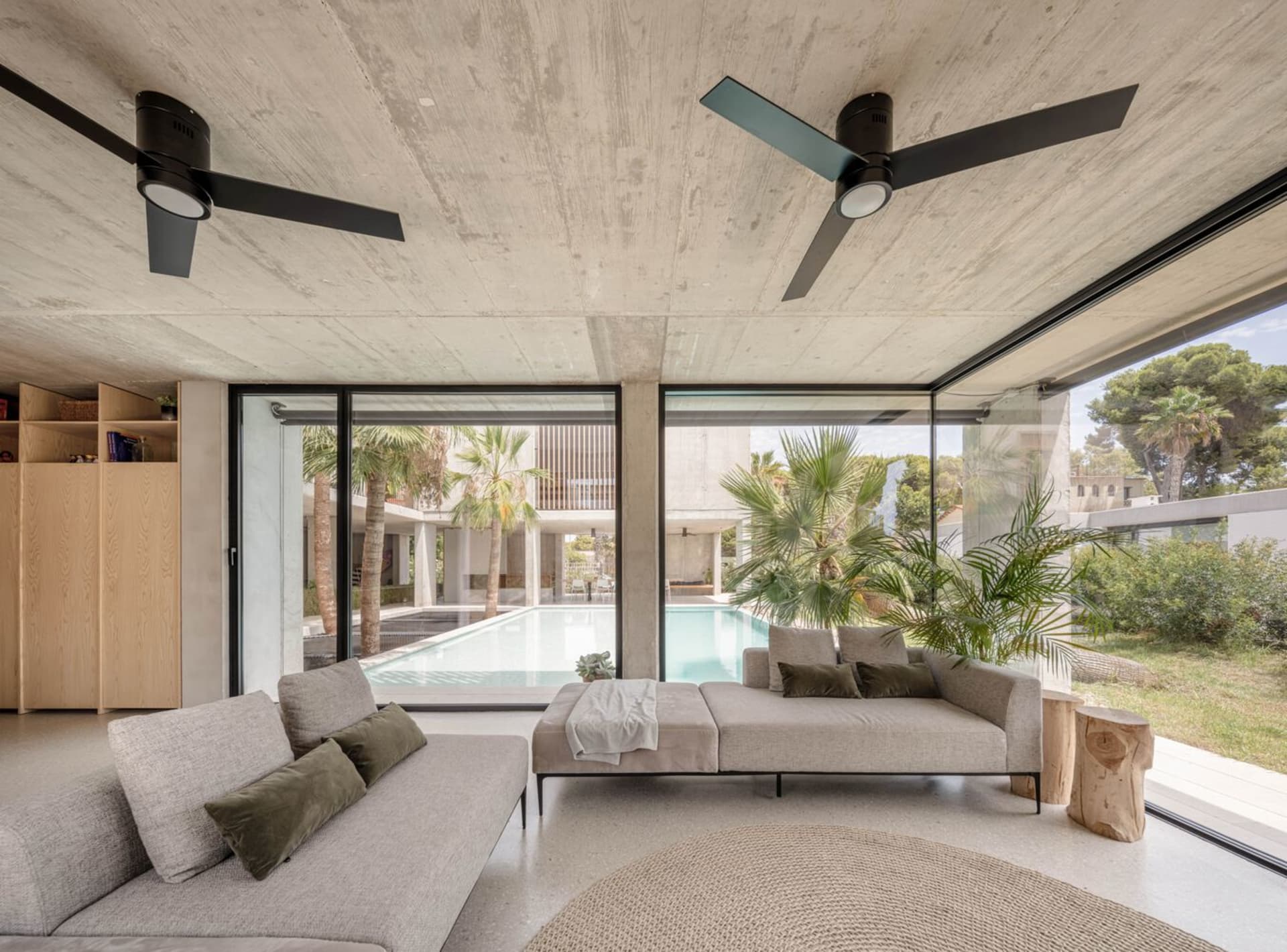 A modern living room featuring a sleek concrete ceiling and walls, with large windows offering a view of a pool and tropical 