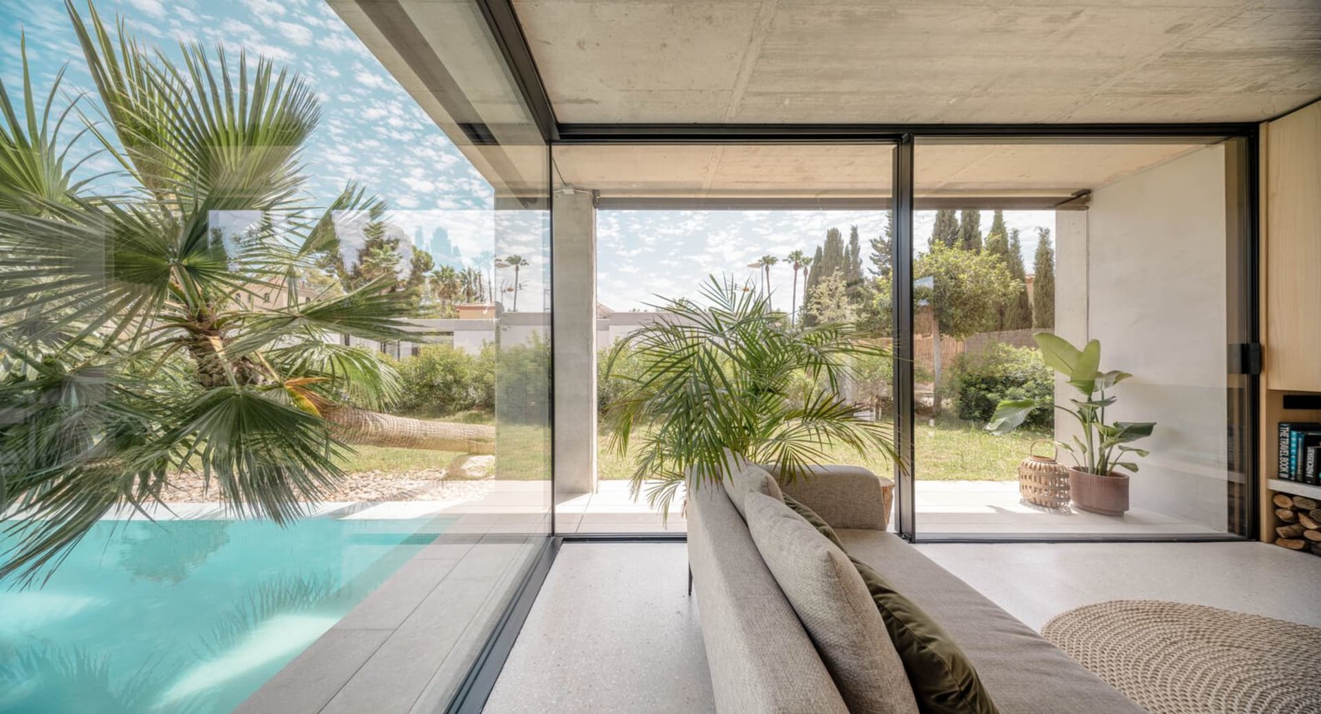 Modern living room with concrete walls and large glass window looking out to an outdoor view of palm trees and a swimming poo