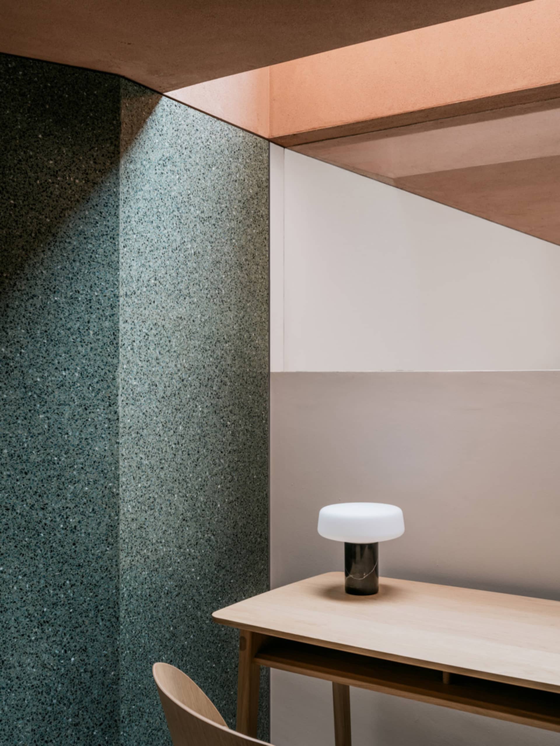 A minimalist office space featuring a desk made of wood with a modern, round lamp and a wall with a combination of terrazzo a