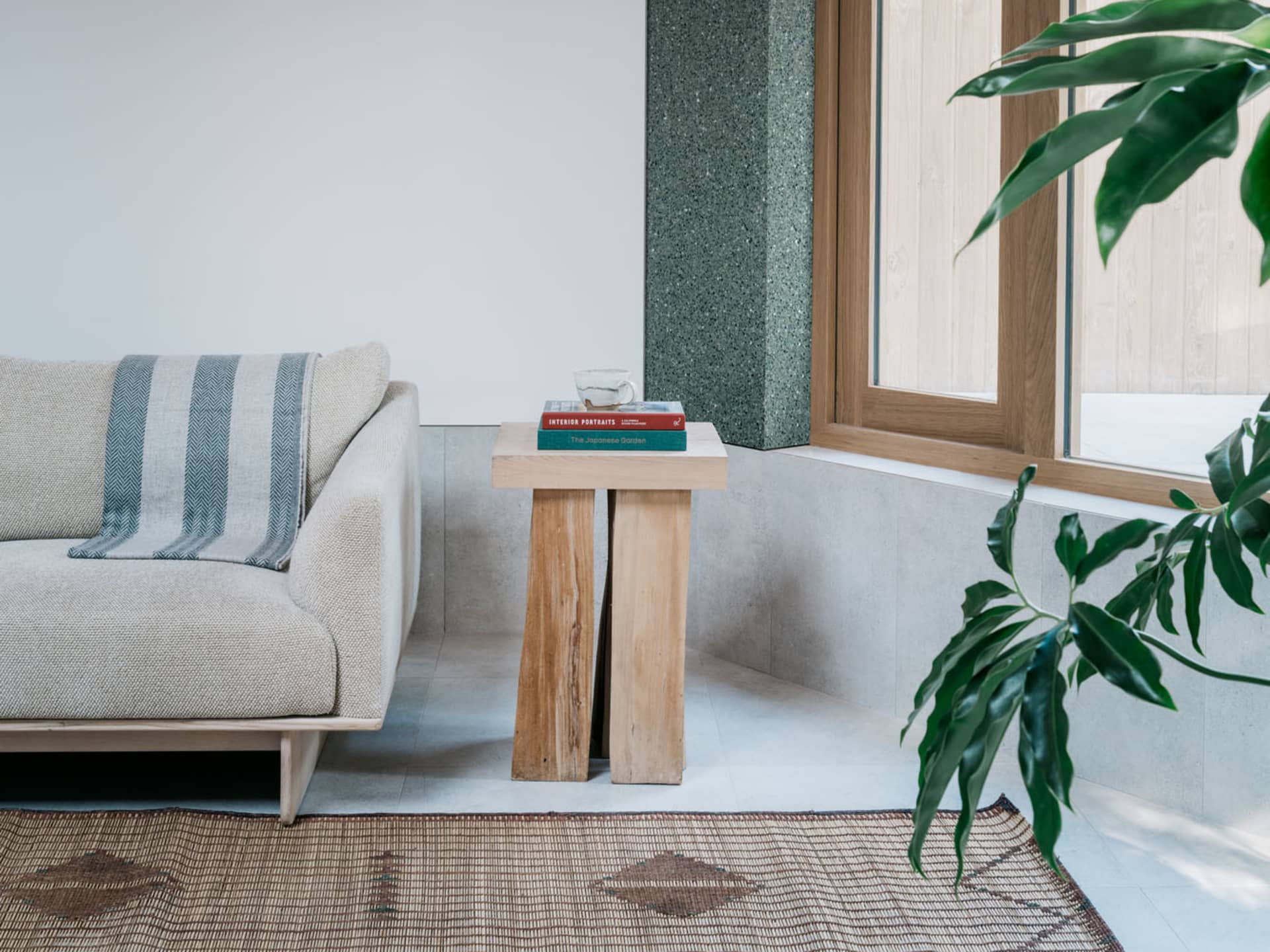 A minimalist living room featuring wooden furniture, a beige sofa, a wooden side table with books and a cup, and a plant in f