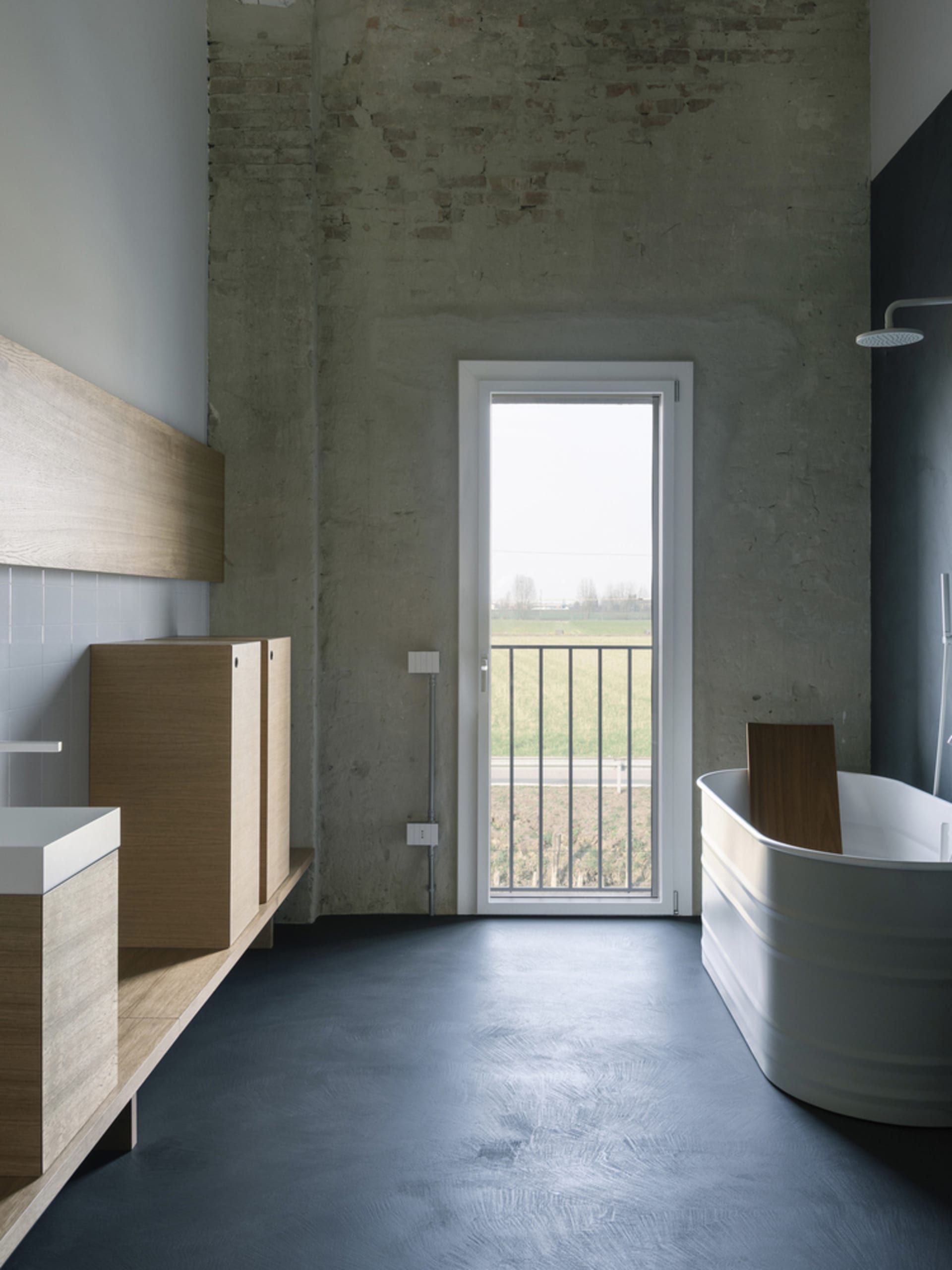Modern bathroom with industrial design featuring concrete floor, wooden vanity, and white bathtub with a view of green fields